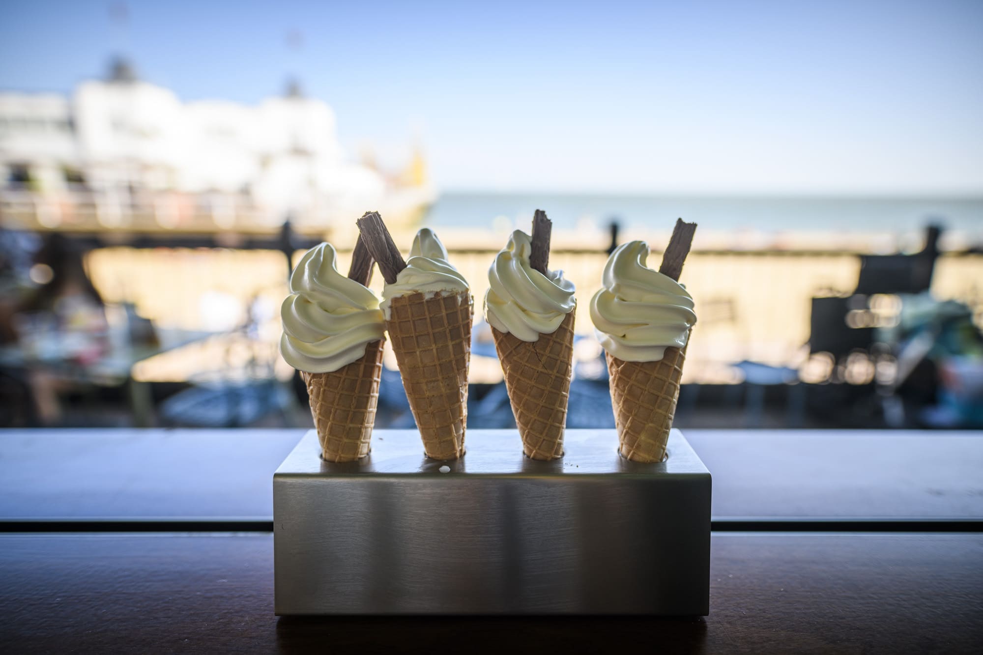 Four Martello soft-serve ice cream cones with flake sticks stand upright in a metal holder, set against a blurred beach and boardwalk background.
