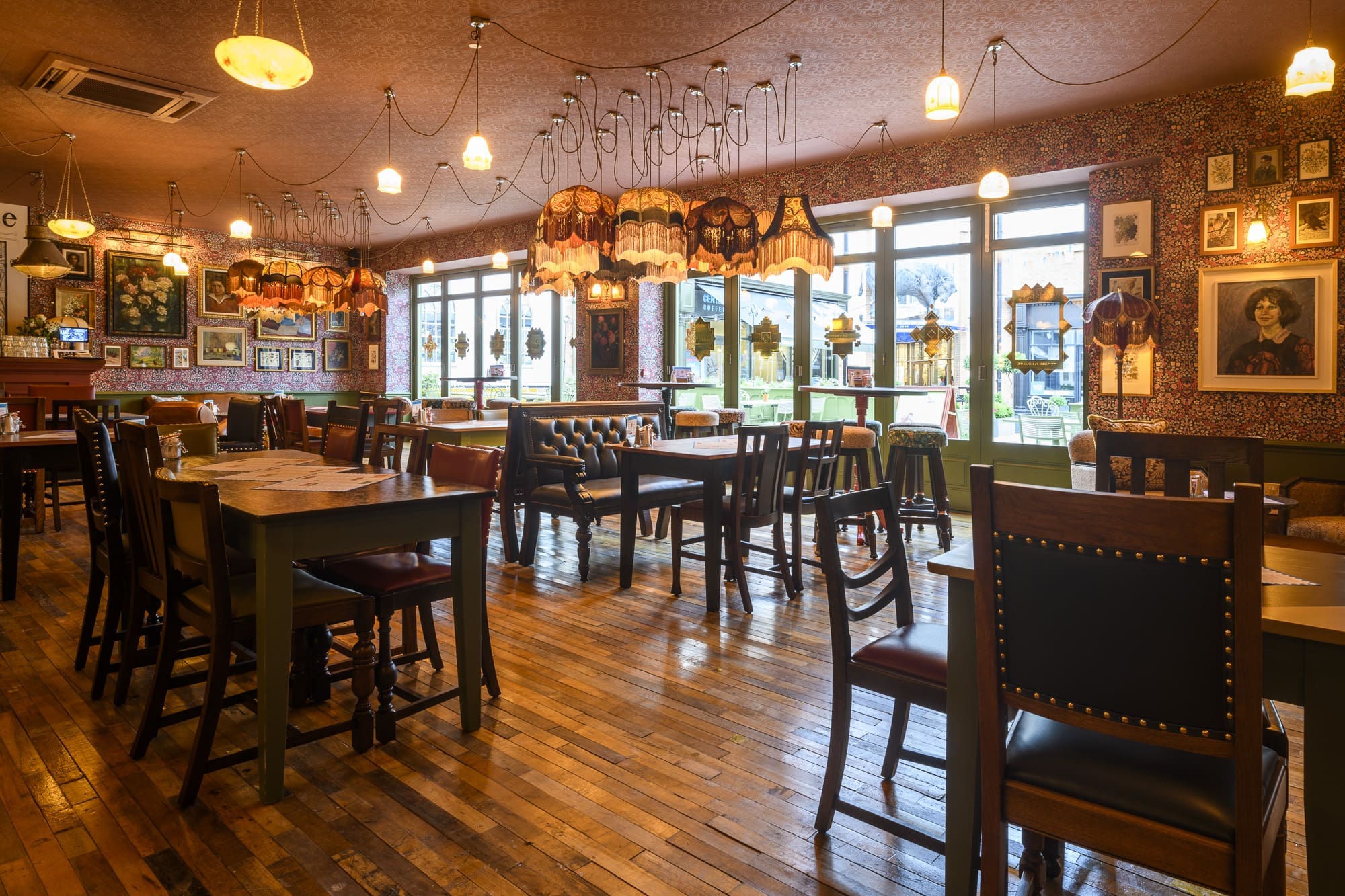 A cozy restaurant, Martino, with wooden floors, vintage lampshades hanging from the ceiling, framed pictures on patterned walls, and large windows letting in natural light. Tables and chairs are arranged throughout the space.