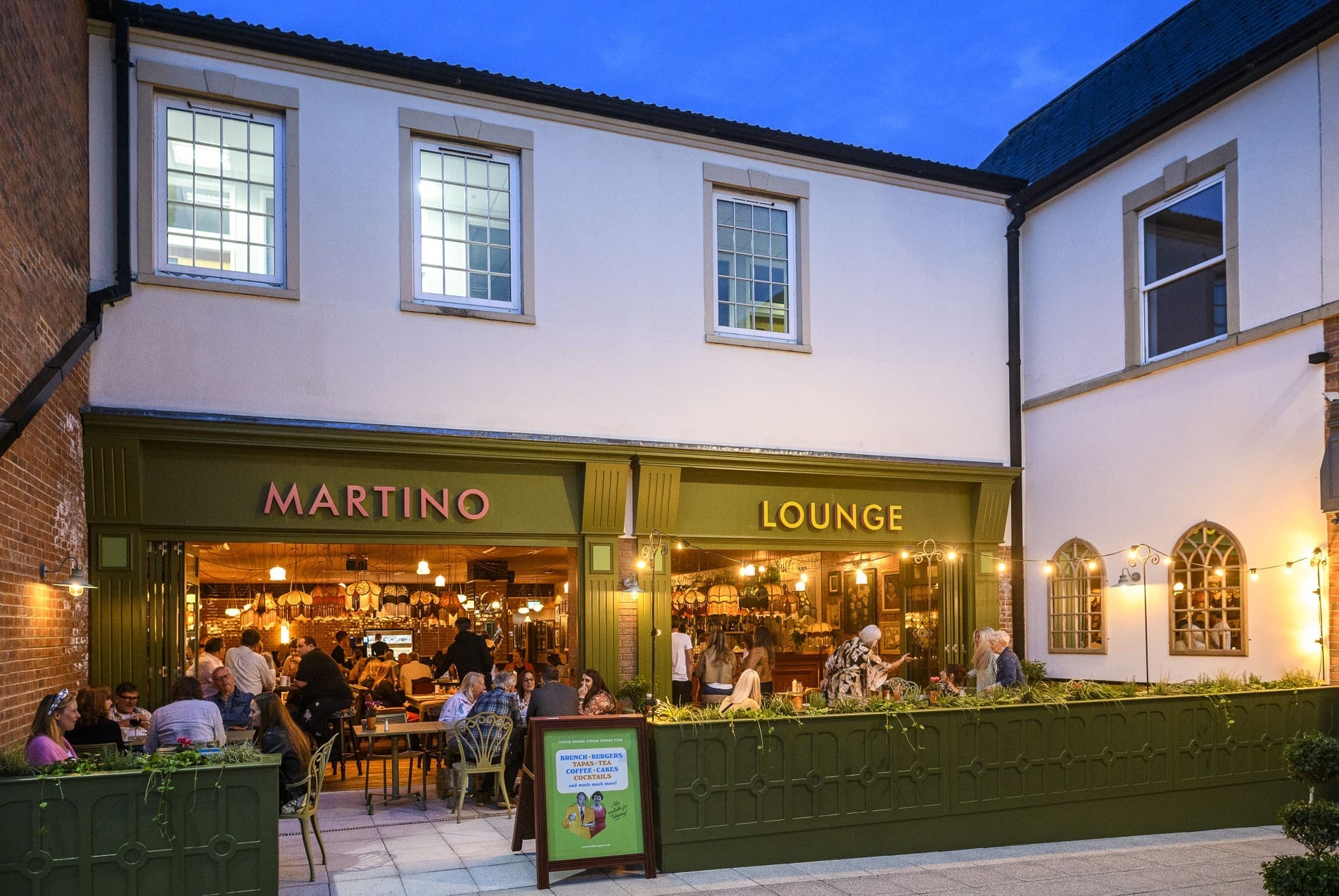 People dine and socialize at outdoor tables in front of Martino Lounge, a vibrant restaurant with green facades and warm lighting. The building, named for its iconic Martino roots, features large windows and decorative string lights.