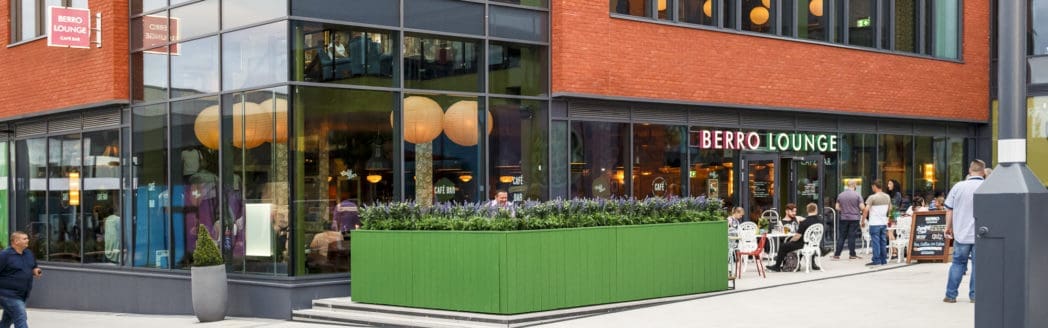 A modern café with large glass windows, red brick walls, and a green planter box outside. People relax at tables on the outdoor patio near the "BERRO LOUNGE" sign, enjoying the vibrant Berro atmosphere as pedestrians stroll by.