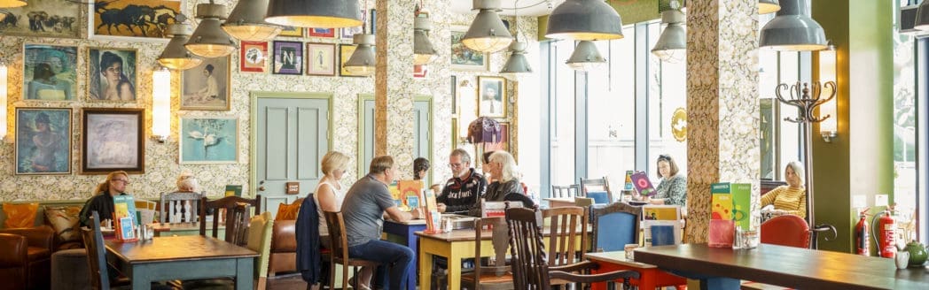 People sit at tables in Berro, a brightly lit, eclectic café with patterned wallpaper, framed art, and vintage decor. Hanging lamps, large windows, and colorful furniture create a cozy, lively atmosphere.