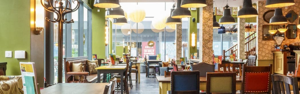 A cozy, brightly lit café with green walls, wooden tables, mismatched chairs, and pendant lights invites you in. Large windows let in natural light, while a staircase and Berro-inspired eclectic decor add unique charm to the space.