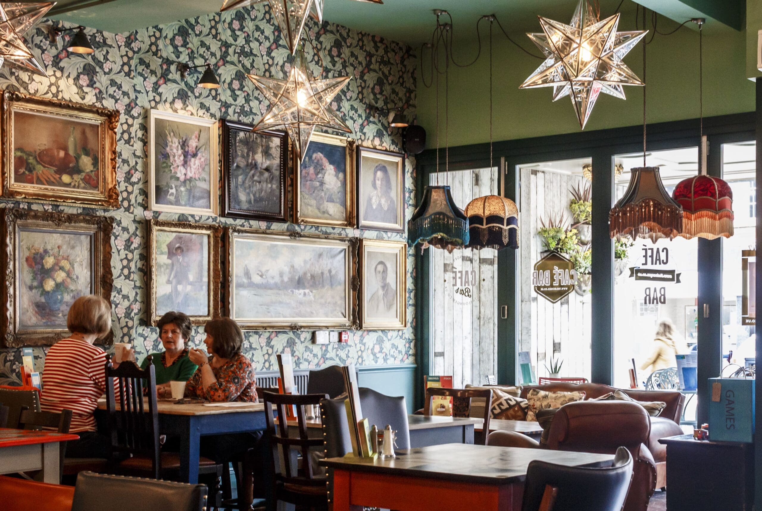 A cozy café with vintage decor by Granello, featuring framed paintings on patterned wallpaper, star-shaped pendant lights, and colorful lampshades. Three women chat at a table as sunlight streams in through the glass doors.