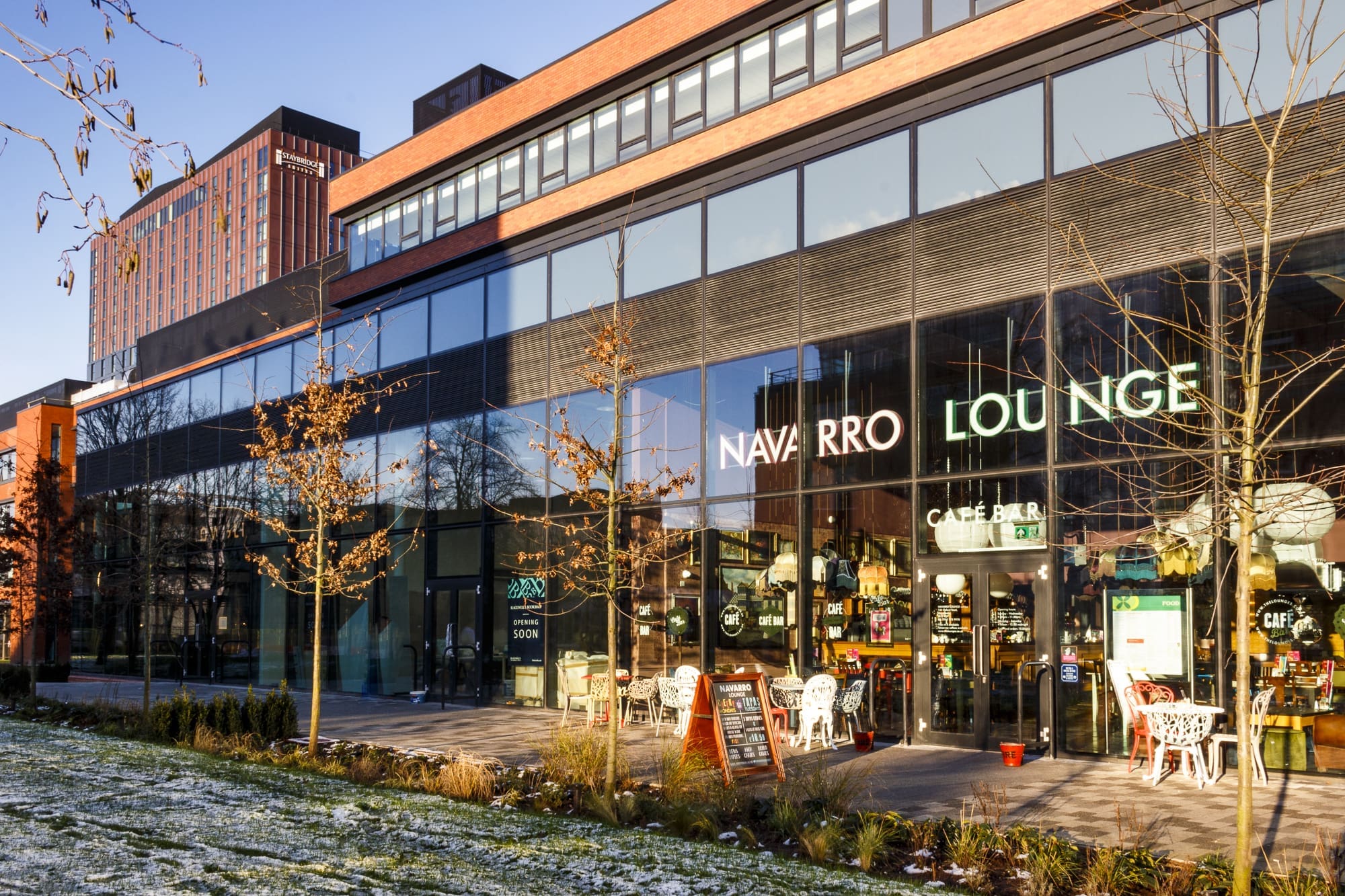 A modern glass-fronted building with prominent "Navarro Lounge" signage. Outdoor tables and chairs line the patio, while trees and patches of snow surround the inviting scene at Navarro’s contemporary setting.
