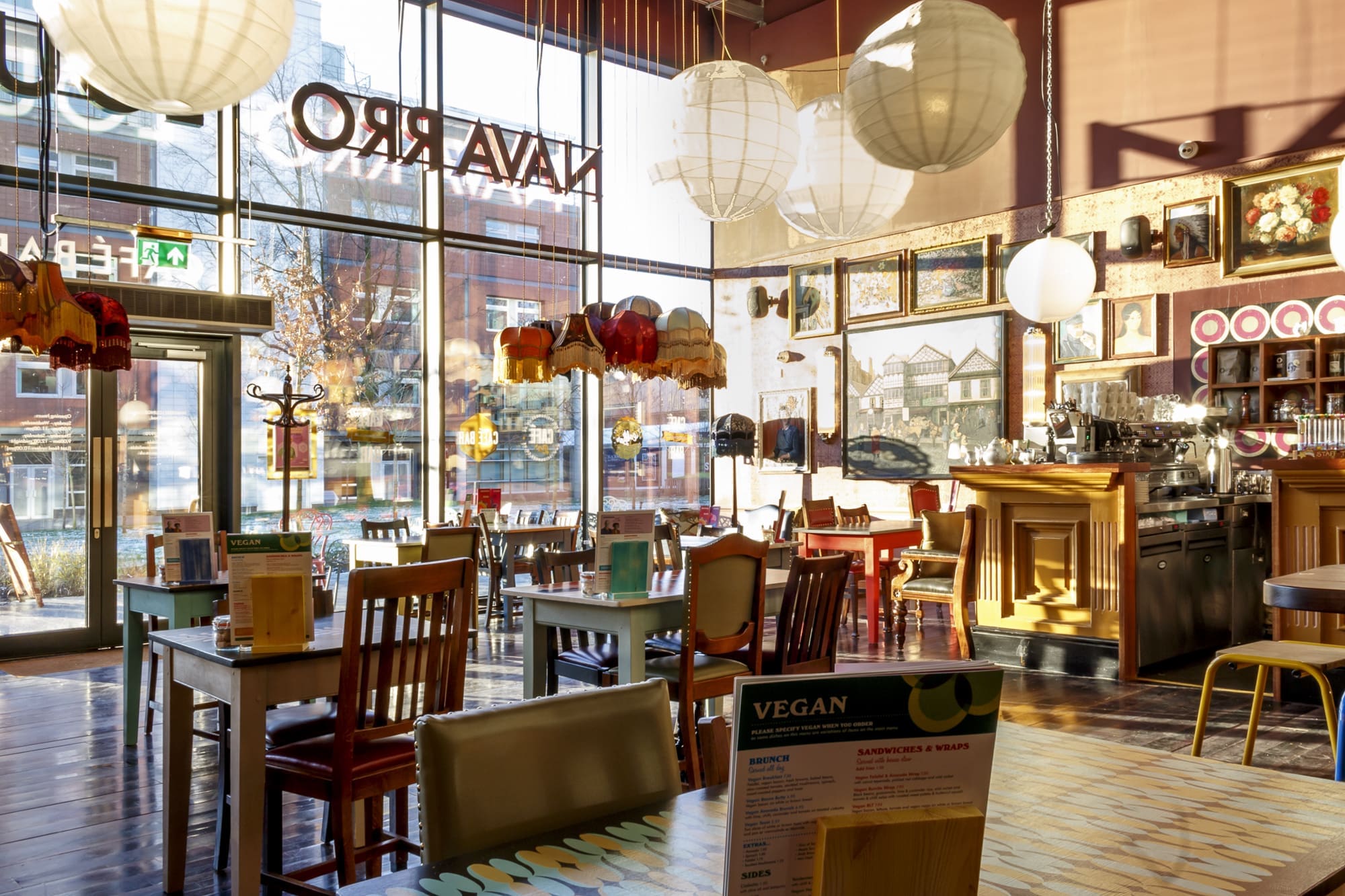 Sunlit Navarro café interior with eclectic furniture, hanging paper lanterns, framed artwork on the walls, and large windows overlooking the street. Tables with menus and a barista area complete this inviting space.