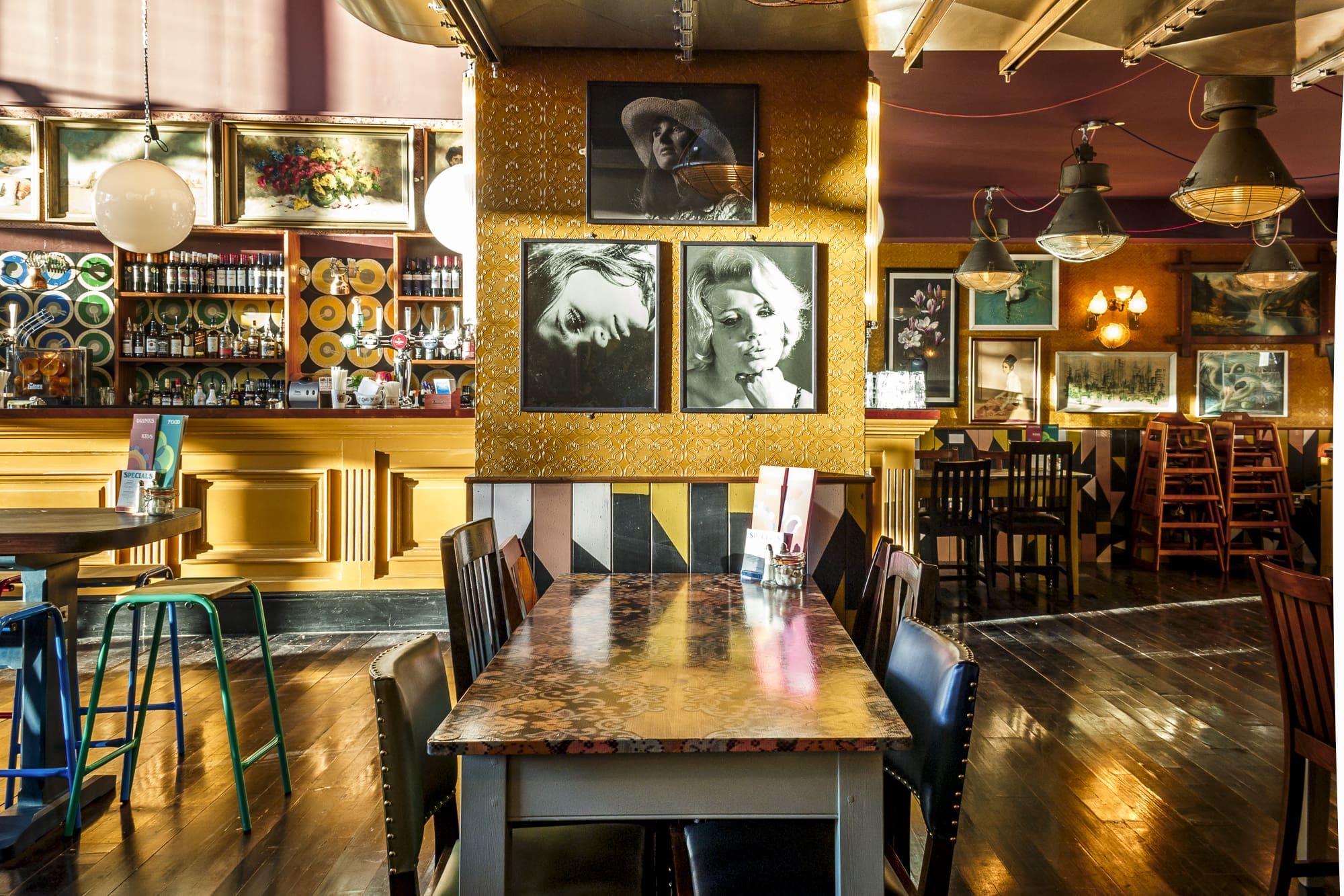 A brightly lit, eclectic restaurant interior with colorful chairs, patterned tables, and hanging lamps. Navarro’s signature touch is seen in the framed black-and-white portraits, decorative plates, and vintage decor adorning the vivid space.