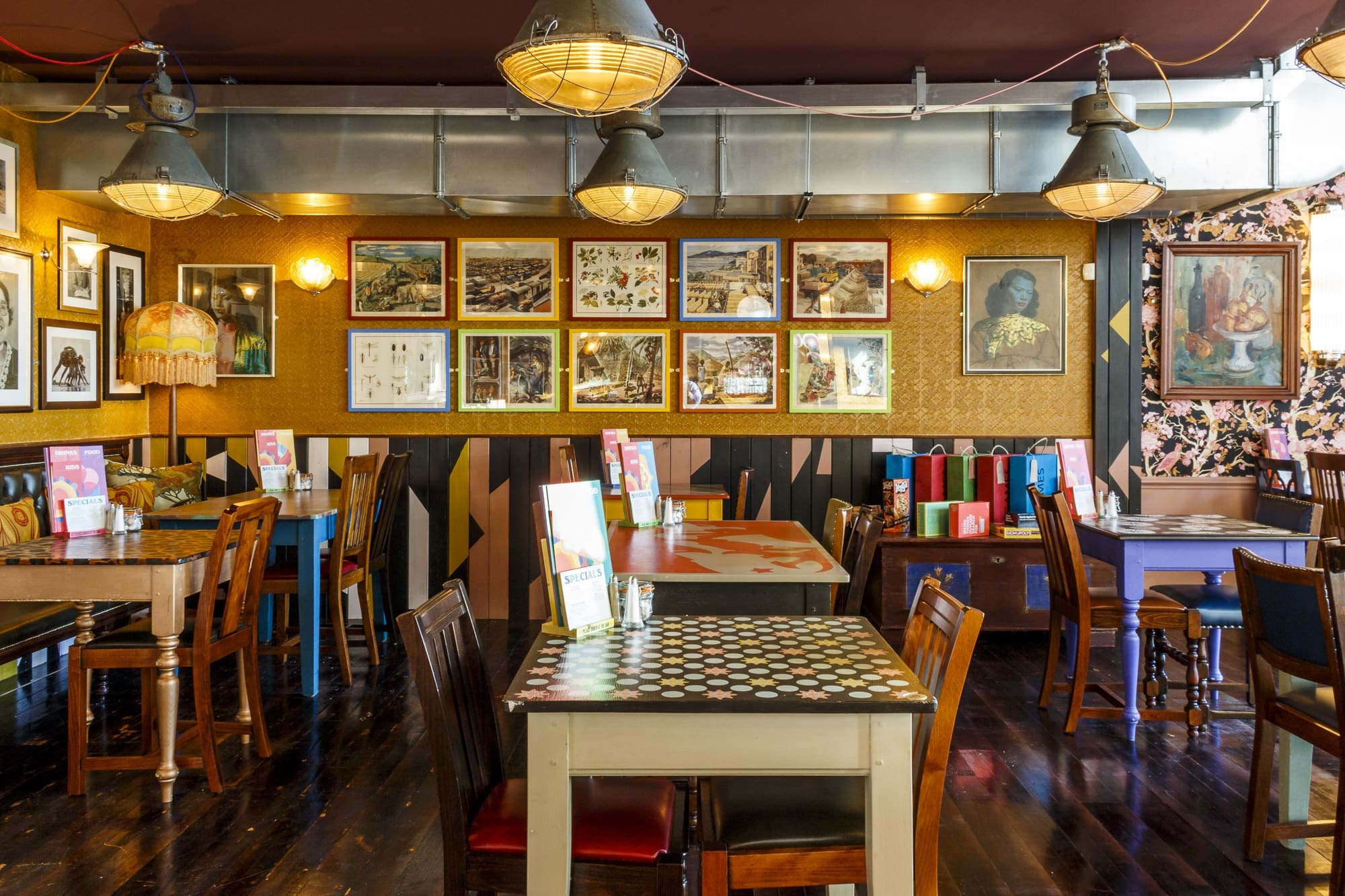 Eclectic Navarro café interior with mismatched tables and chairs, vintage lamps, framed art and photos on a patterned wall, polished dark wood floors, and colorful menus on each table. The space feels cozy and artistic.