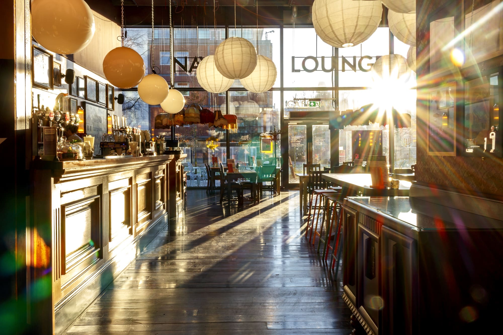 A cozy bar with hanging paper lanterns, sunlight streaming through large windows, casting reflections on Navarro’s polished wooden floor and counters; empty chairs line the bar and tables.