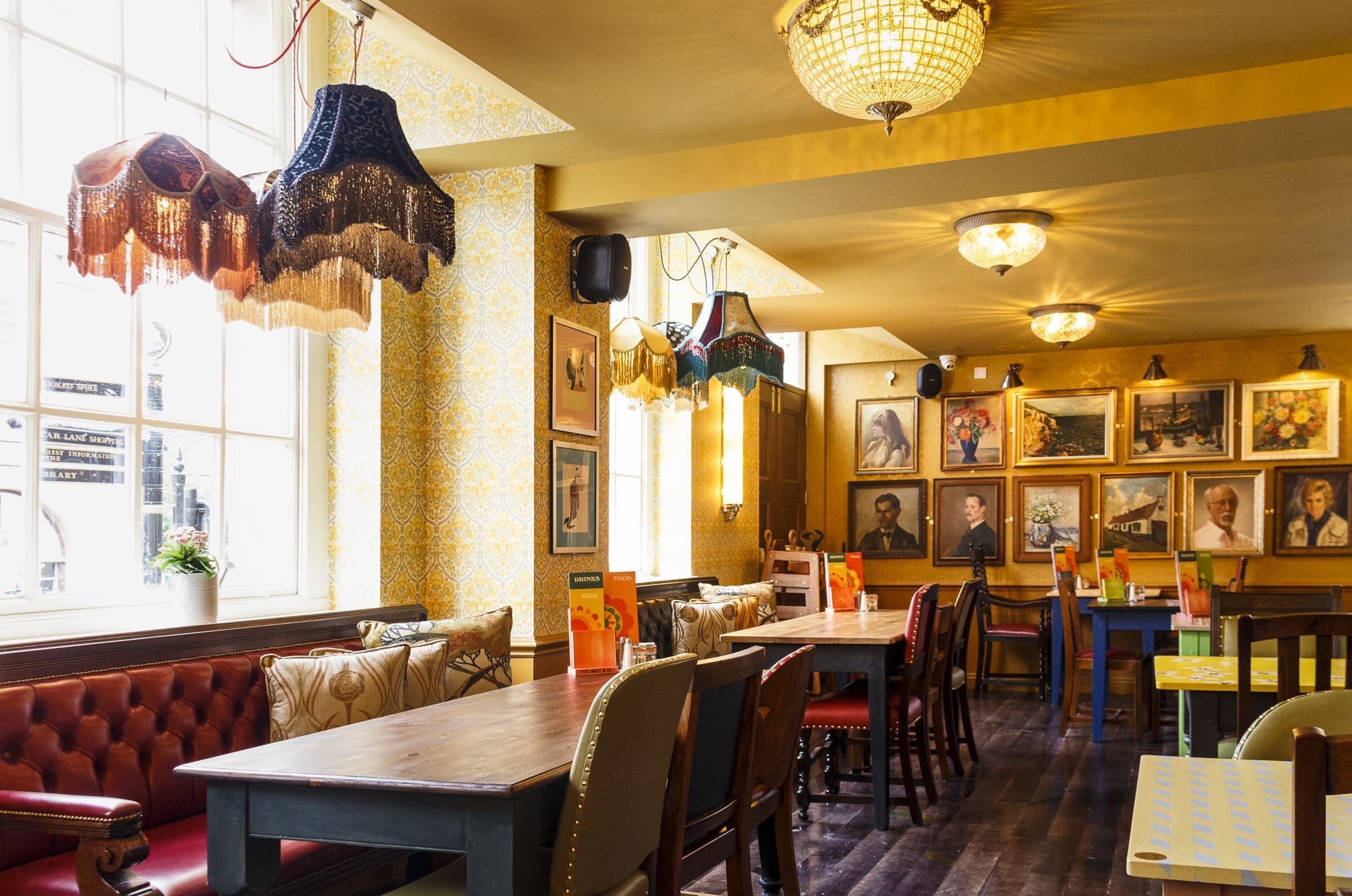 A cozy, eclectic cafe interior with colorful fringed lamps, mismatched chairs, art-covered walls, and tufted red banquette seating beside large sunny windows—perfect for enjoying a Sorbo in style.