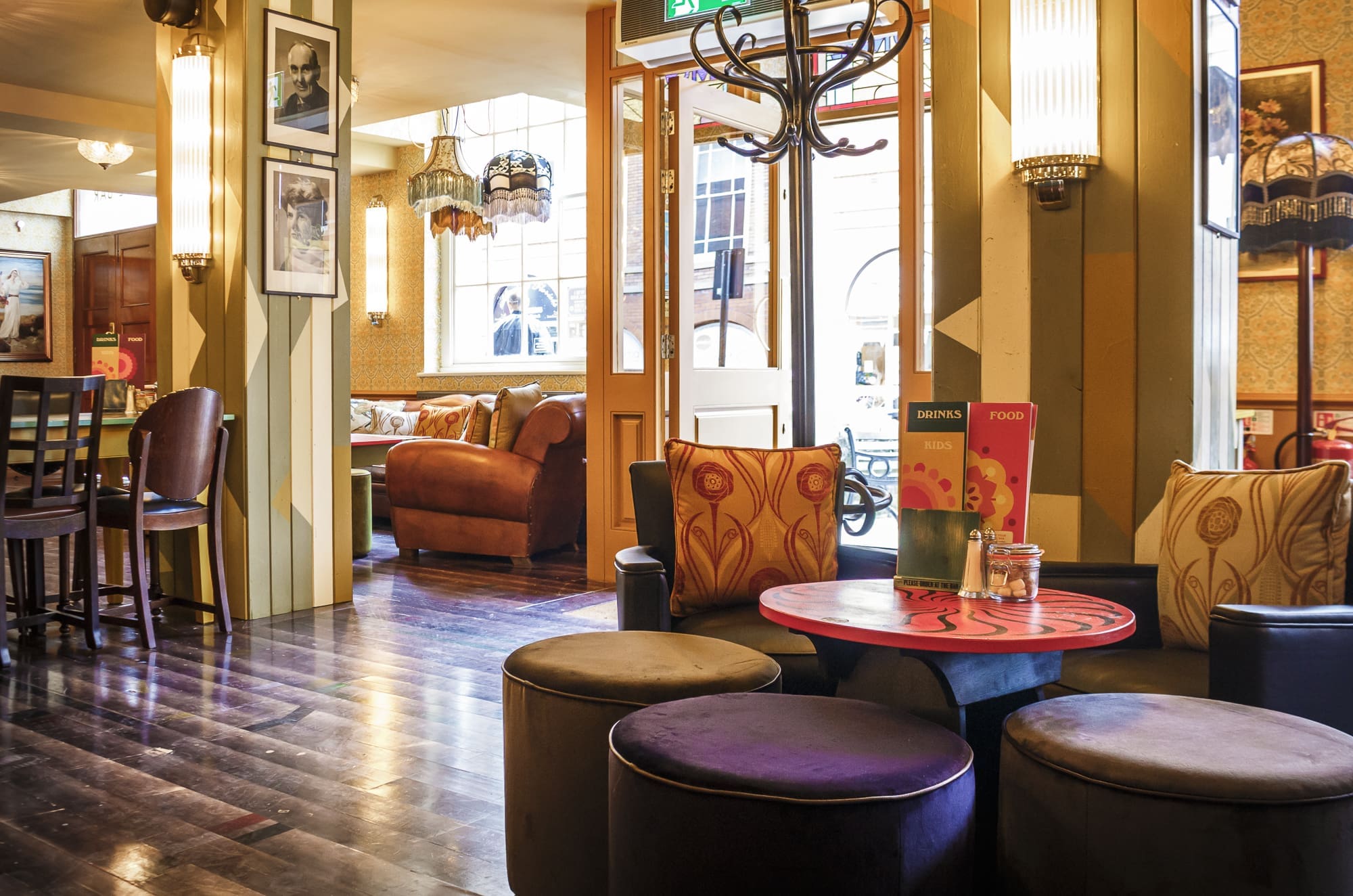 Cozy Sorbo cafe interior with round stools around a small red table, plush armchairs, framed photos on the walls, hanging lamps, and large windows letting in natural light.