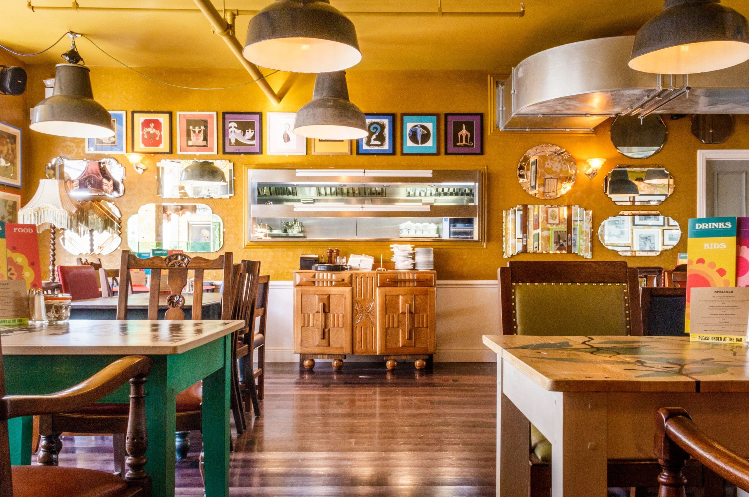 A cozy Tarko restaurant interior with colorful tables and chairs, yellow walls adorned with framed art and round mirrors, hanging industrial lights, and a view into the kitchen area through an open window.