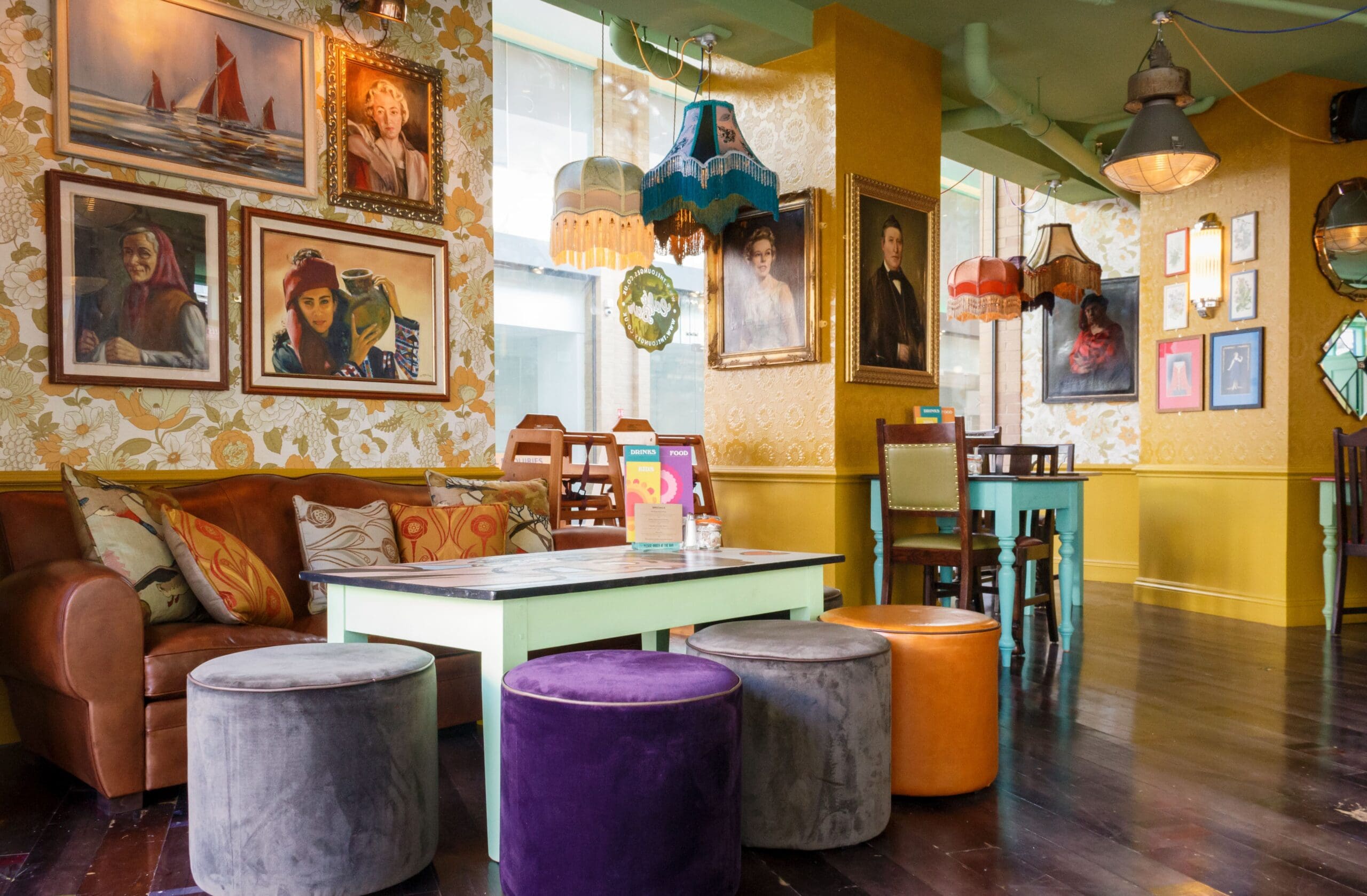 A colorful, eclectic café interior by Tarko features mismatched furniture, patterned wallpaper, vintage portraits, hanging lampshades, and plush stools around a green coffee table. Large windows let in natural light.