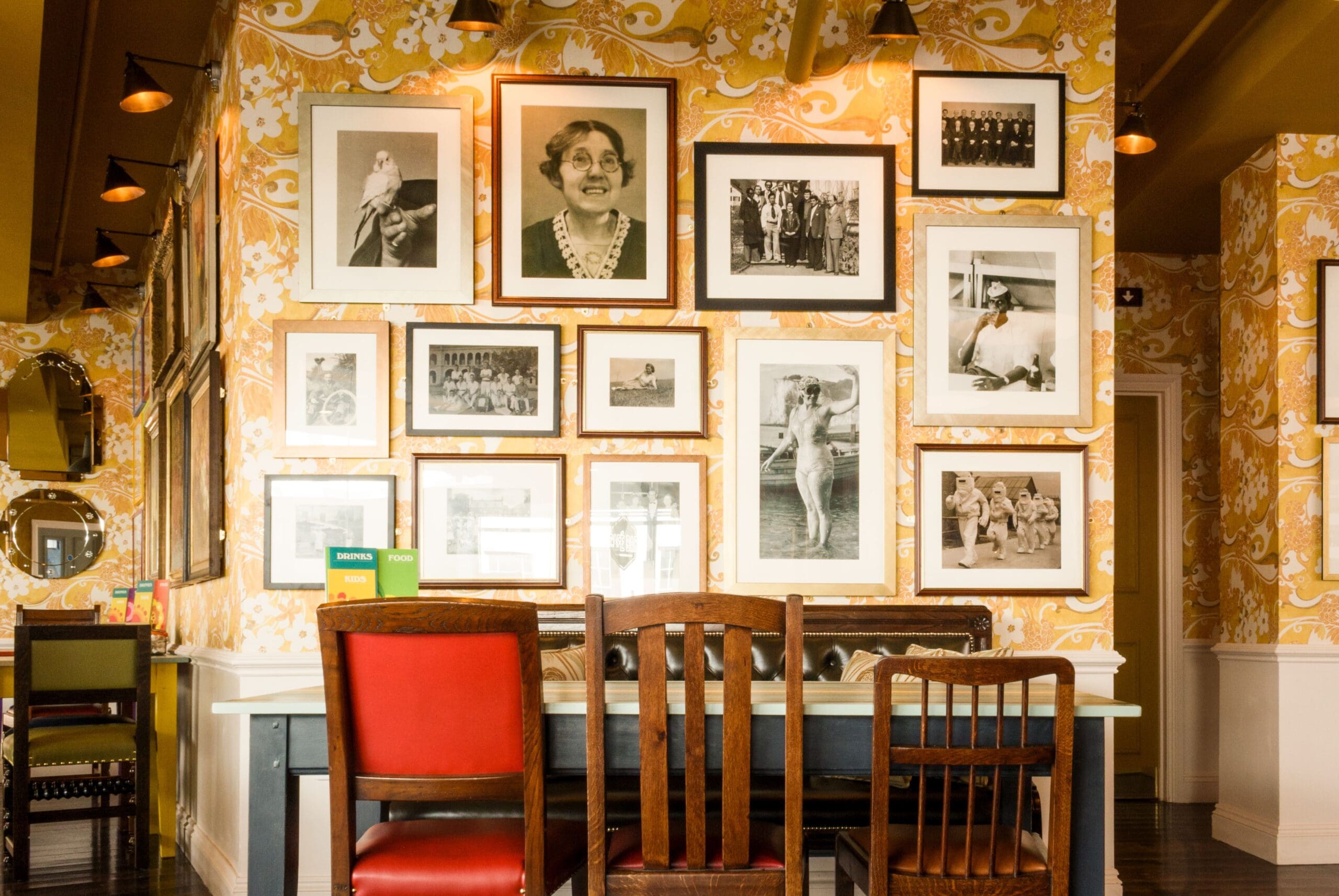 A dining area with a long Tarko table and mismatched chairs sits against a wall decorated with vintage black-and-white photographs and floral yellow wallpaper. Warm lighting creates a cozy, eclectic atmosphere.