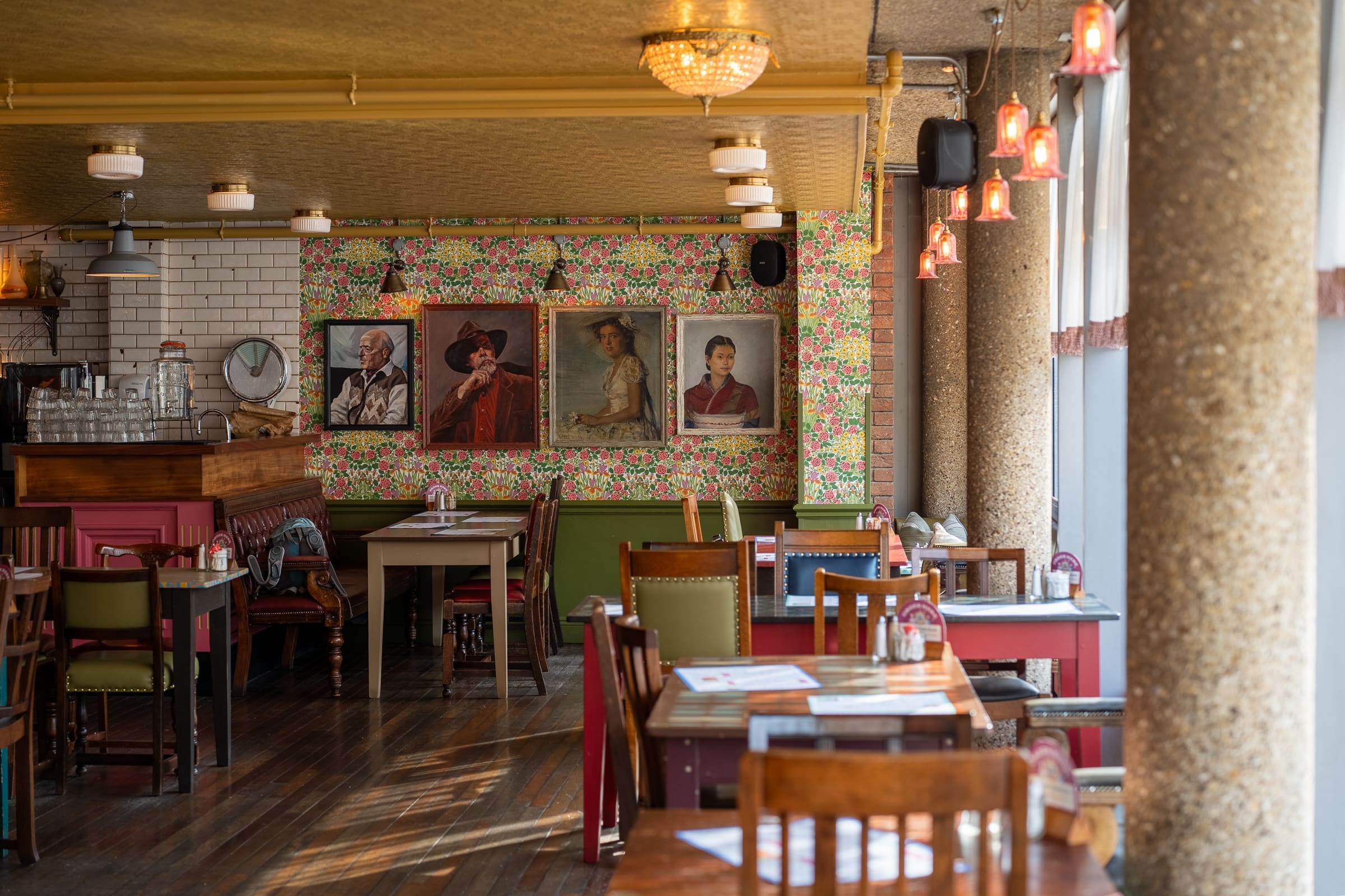 A cozy, sunlit Mercado restaurant interior with wooden tables and chairs, floral wallpaper, and four framed portraits on the wall. Soft pendant lights hang from the ceiling, and large windows let in natural light.
