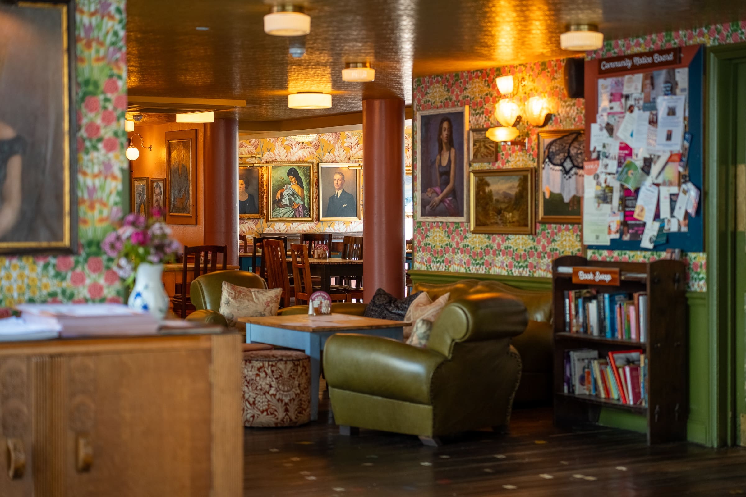 Cozy interior of a cafe or lounge inspired by Mercado style, with green armchairs, floral wallpaper, framed portraits, bookshelves, and a community notice board. Warm lighting creates a welcoming atmosphere.