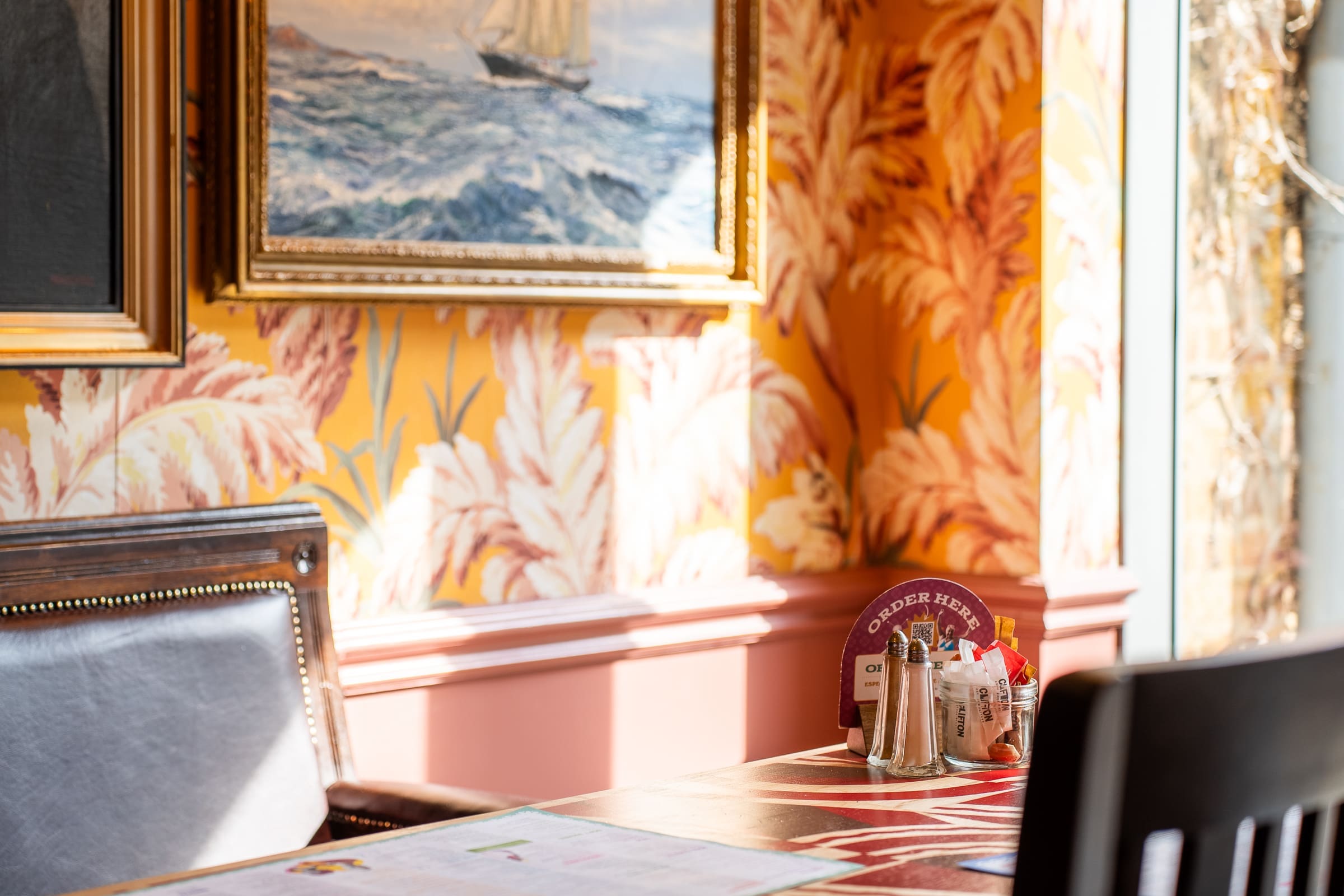 A sunlit Mercado restaurant table with condiments and menus sits by a window. The walls feature bold yellow wallpaper with pink leafy patterns and framed paintings, including one of a sailboat on the ocean.