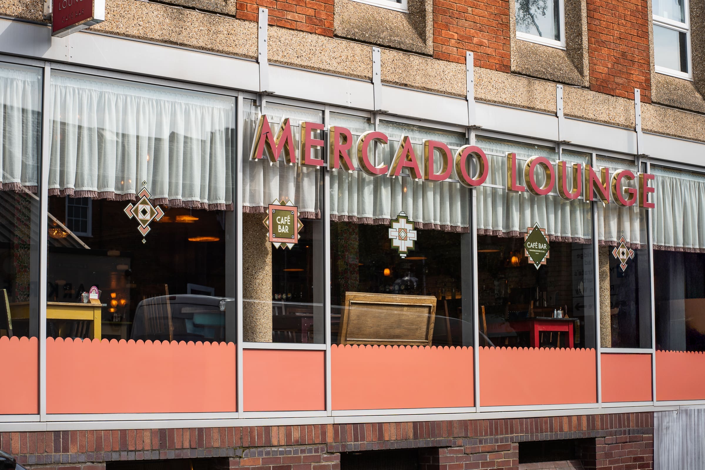 Street view of the Mercado Lounge café, featuring large windows with red Mercado lettering, decorative signs, white curtains, and a glimpse of tables and chairs inside. The building showcases a red brick and concrete exterior.