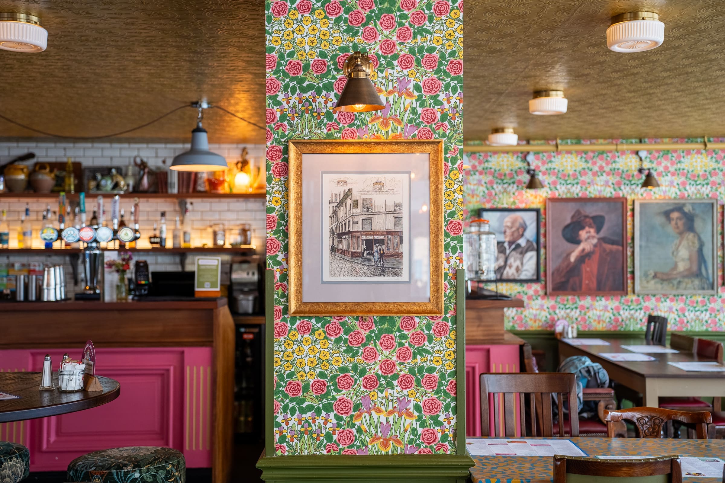 A vibrant pub interior with floral wallpaper, framed artwork, and wooden furniture. The bar area has bottles and taps, reminiscent of a Mercado, while walls display portraits and a street scene drawing. Warm lighting creates a cozy atmosphere.