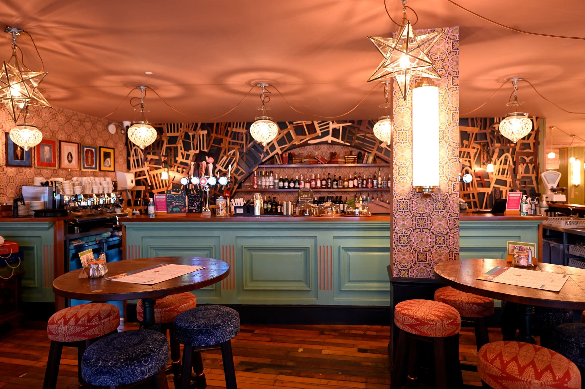 A colorful bar with green paneling, patterned walls, star-shaped pendant lights, round tables with Merletto-patterned stools, and various bottles and glasses displayed behind the counter. The atmosphere is cozy and eclectic.