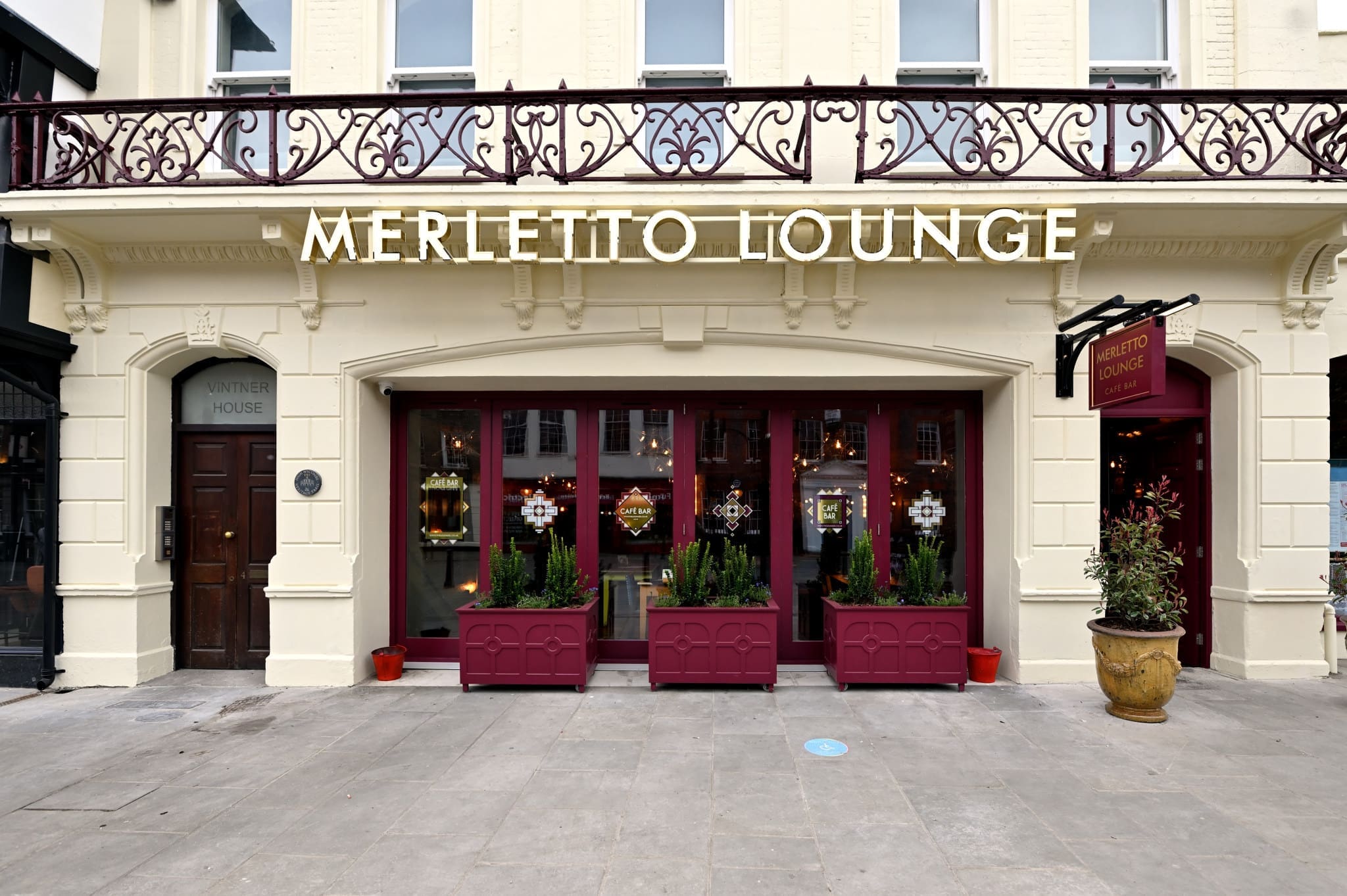 The exterior of Merletto Lounge, a cream-colored building with burgundy-framed windows, door, and flower planters, welcomes guests. Warm lights and decor glow inside Merletto, with its distinctive sign hanging above the entrance.