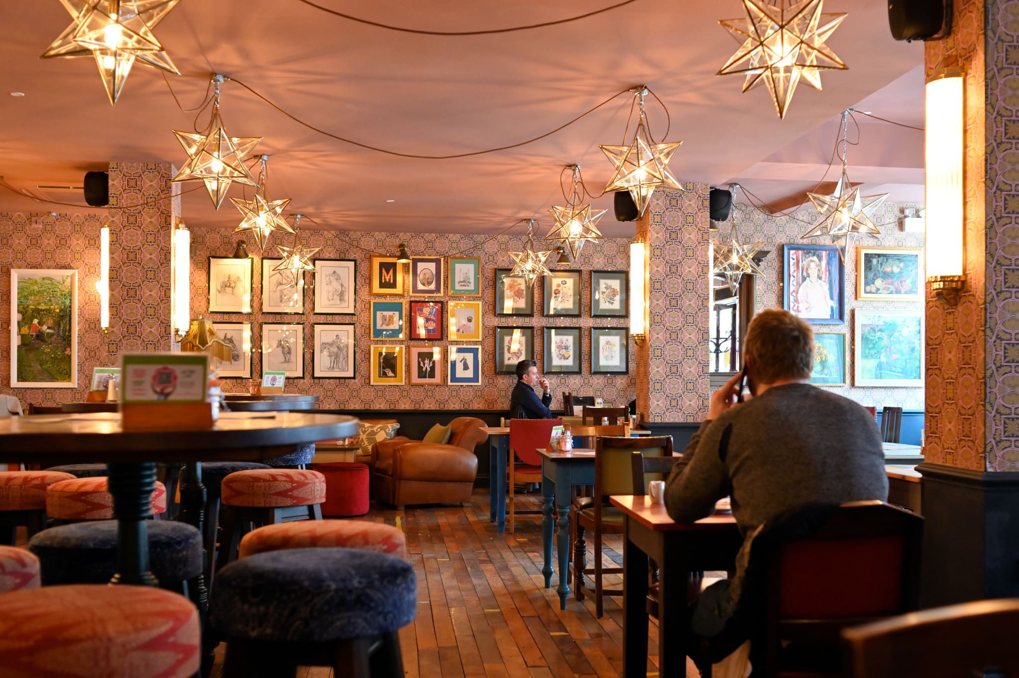 A cozy café or restaurant with star-shaped pendant lights, patterned Merletto-inspired walls adorned with framed art, wooden floors, and a few people sitting at tables. The atmosphere is warm and inviting.