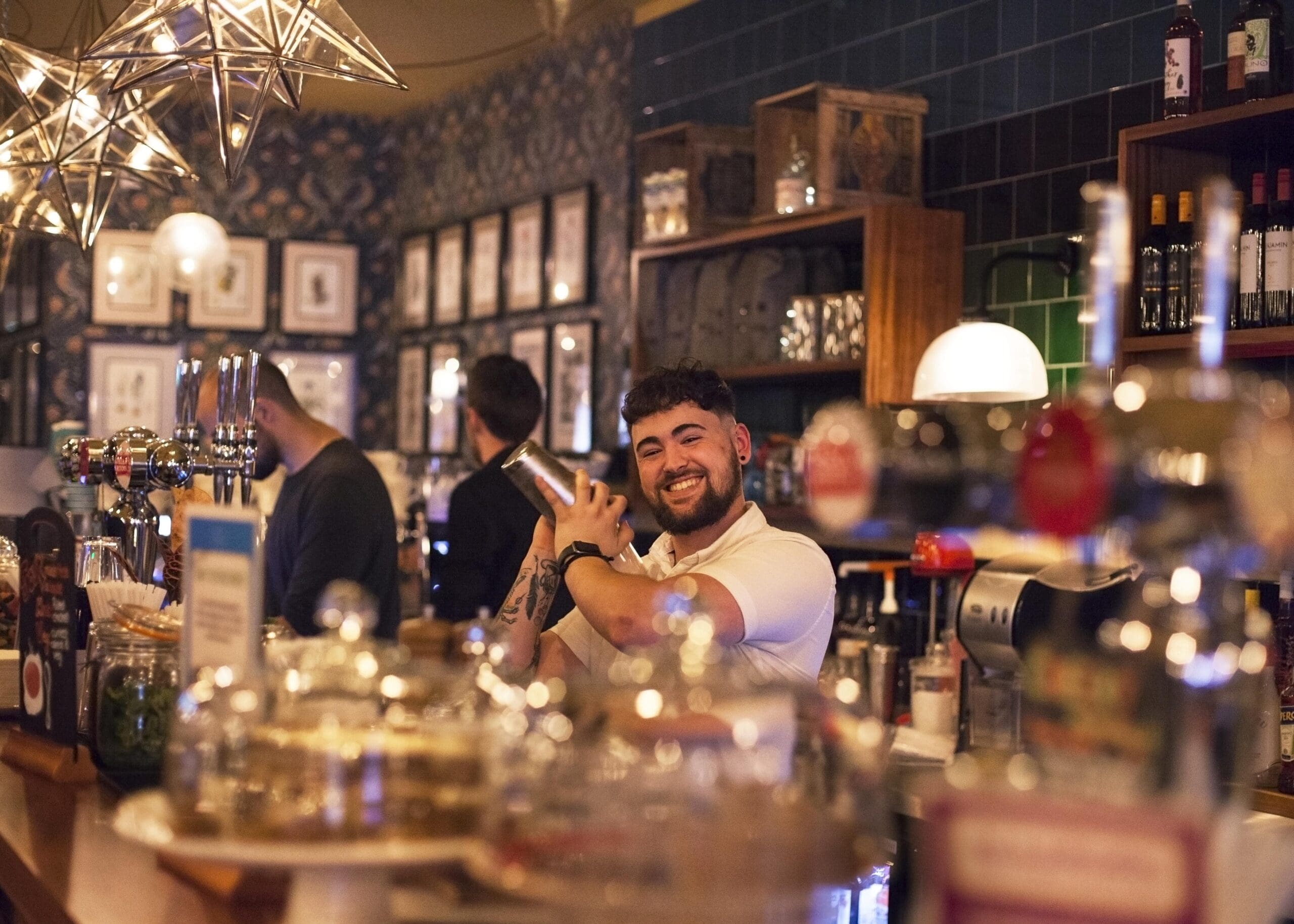 A smiling Poco bartender shakes a cocktail shaker behind the bar, surrounded by bottles, glasses, and decorative lights, with patrons and framed pictures in the background.