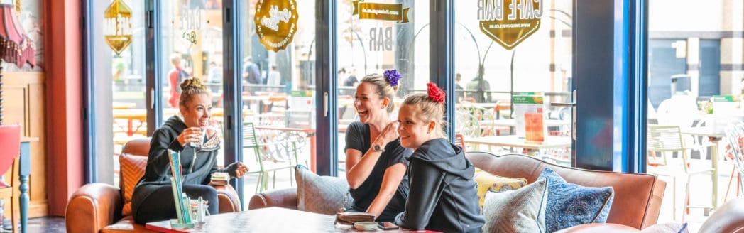 Three women in casual clothing sit on sofas around a coffee table in a bright, modern Casco cafe, smiling and laughing together. Large windows reveal outdoor seating and pedestrians outside.