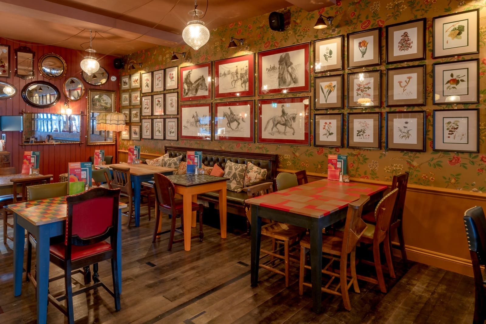 Cozy café with mismatched tables and chairs, floral wallpaper, assorted framed artwork on the walls, and menu cards on each table. Warm, cobrizo lighting creates a welcoming atmosphere.