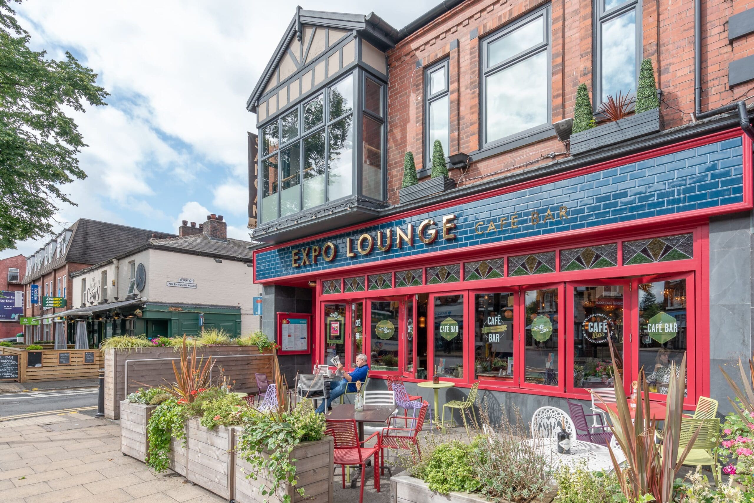 Expo Lounge is a vibrant cafe with striking blue tiles, red trim, and large windows. Colorful outdoor seating and potted plants line the sidewalk, with a person enjoying the lively Expo atmosphere amid nearby buildings and trees.