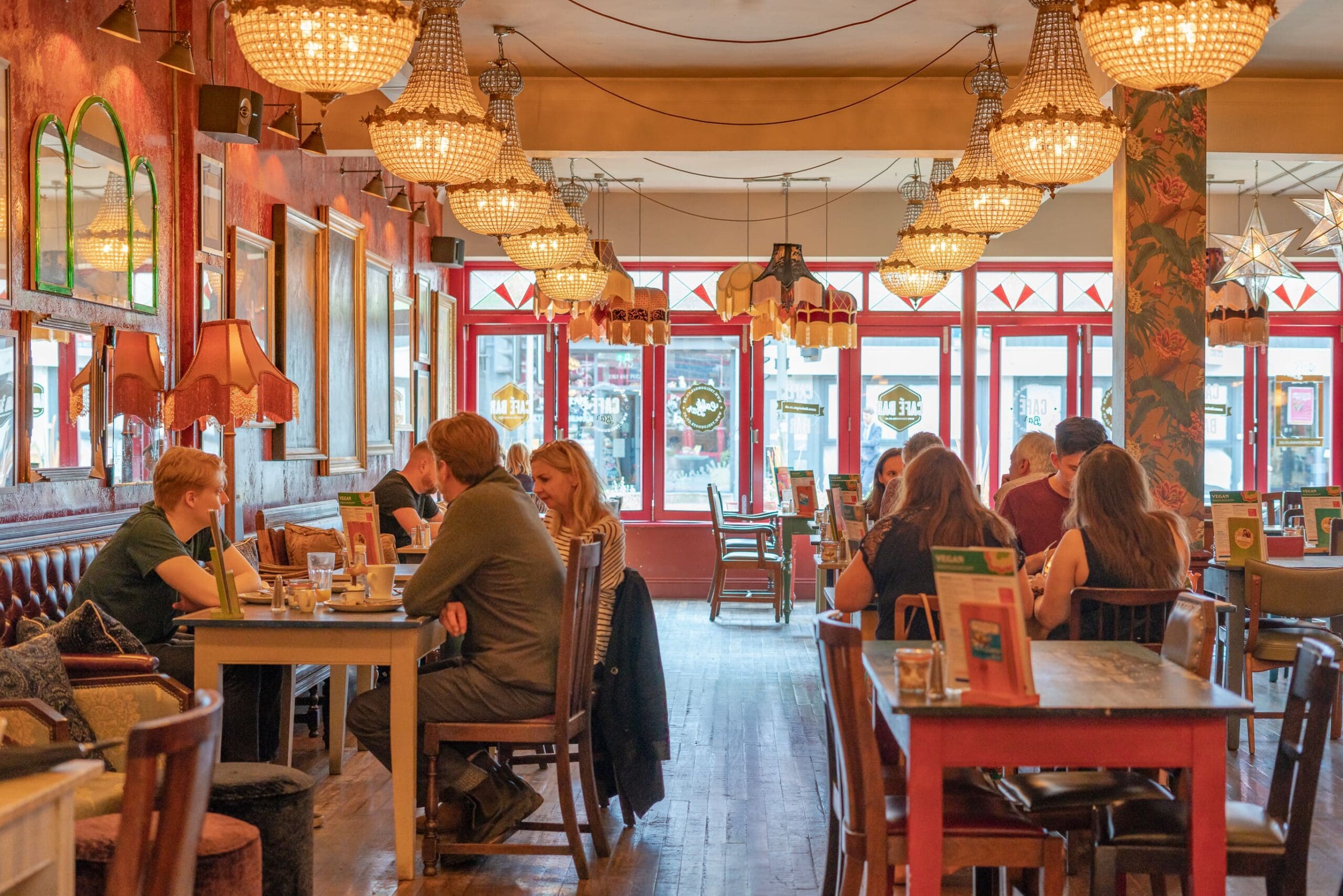 People are dining and talking inside a cozy, warmly lit restaurant, reminiscent of an Expo gathering, with chandeliers, red walls, decorative lamps, and framed art. The inviting atmosphere features several tables occupied by guests.