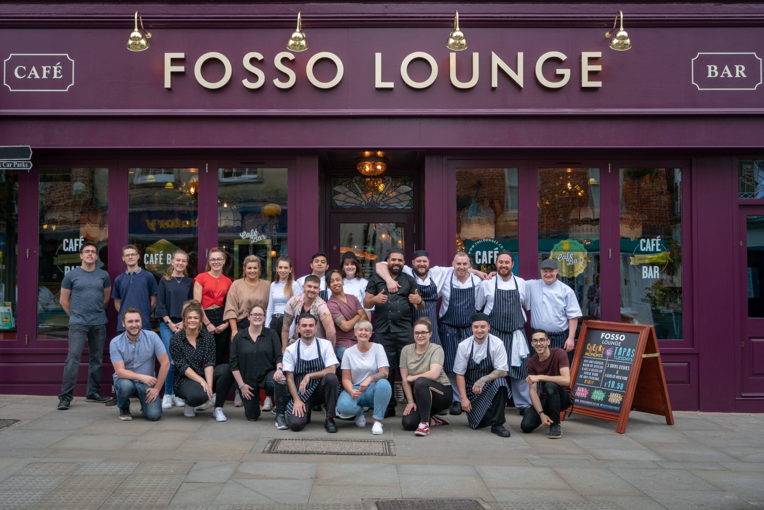 A large group of people pose and smile outside the entrance of Fosso Lounge café bar, standing and kneeling on the sidewalk in front of the fosso purple storefront. A menu board stands near the door.