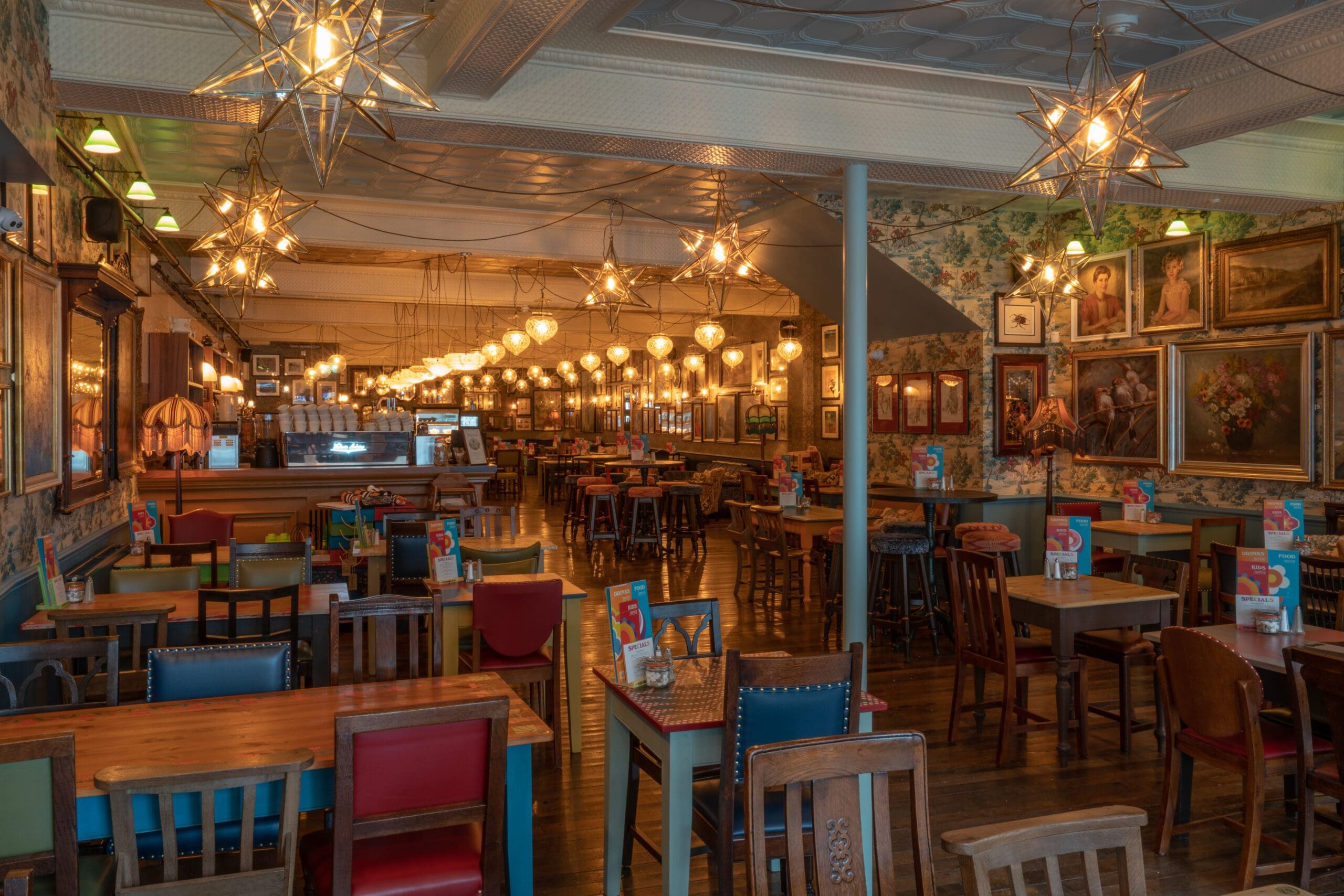 A warmly lit, eclectic restaurant with colorful chairs, wooden tables, star-shaped pendant lights, and walls adorned with framed art and photos creates a fosso of charm. The space is empty yet inviting.