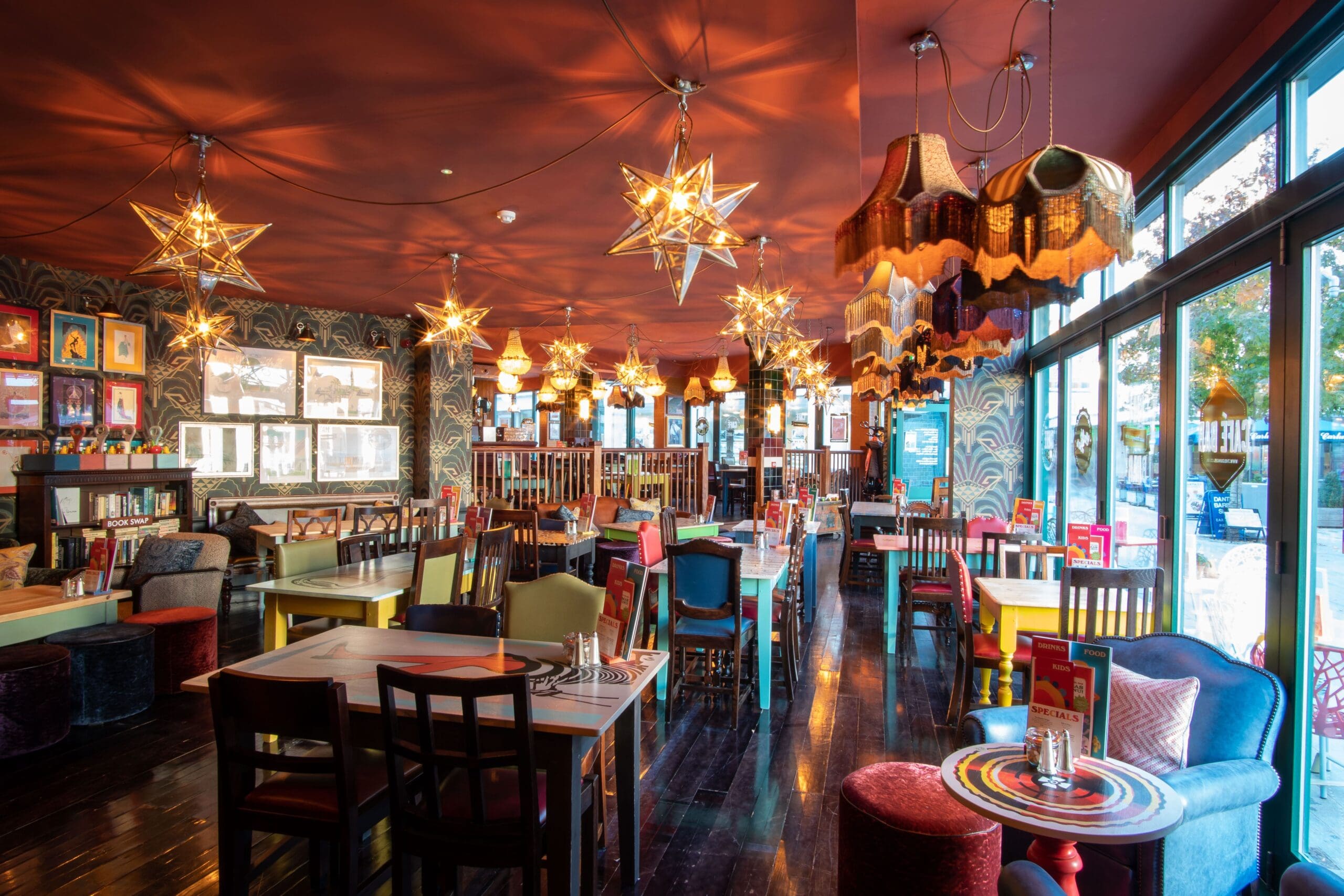 A vibrant restaurant interior features colorful mismatched chairs and tables, Marciano star-shaped pendant lights, retro lampshades, large windows, and eclectic decor with framed art on patterned walls. Sunlight streams in through the windows.