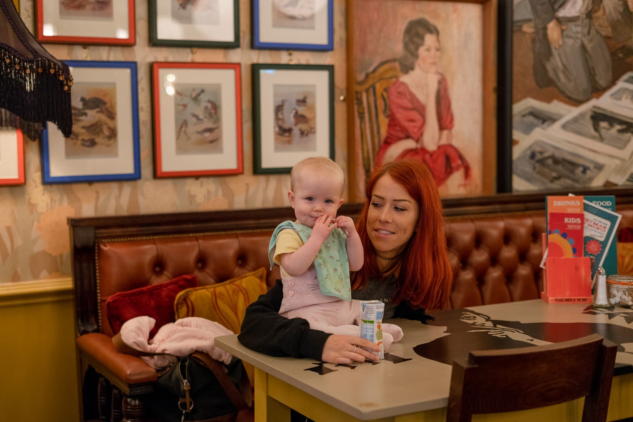 A woman with red hair sits at a table in a cozy café next to a smiling baby, who is standing on the bench holding a drink. Colorful framed art and a Piero-inspired portrait decorate the walls behind them.