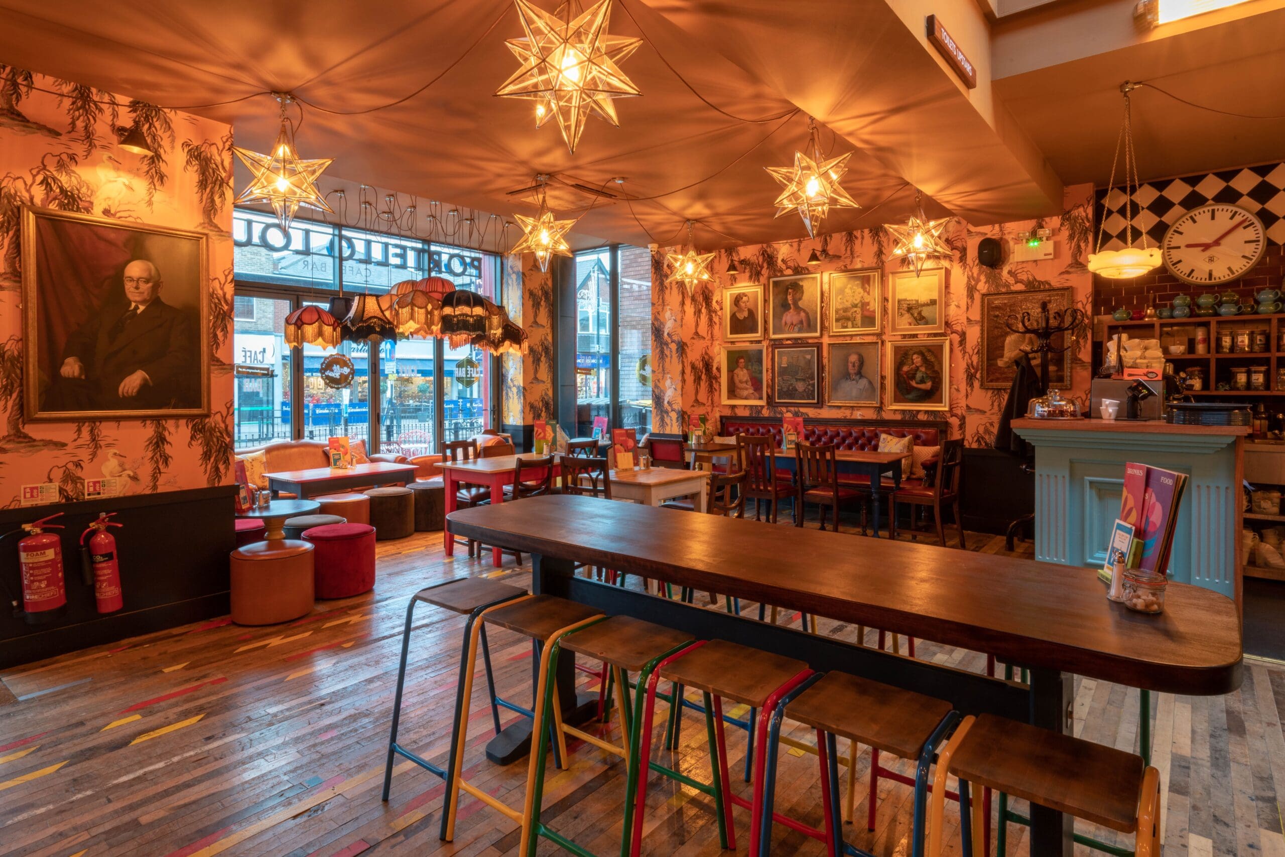 A cozy Portello café interior with warm lighting, eclectic wall art, star-shaped hanging lights, wooden tables and stools, colorful decor, and large windows looking out onto a street.