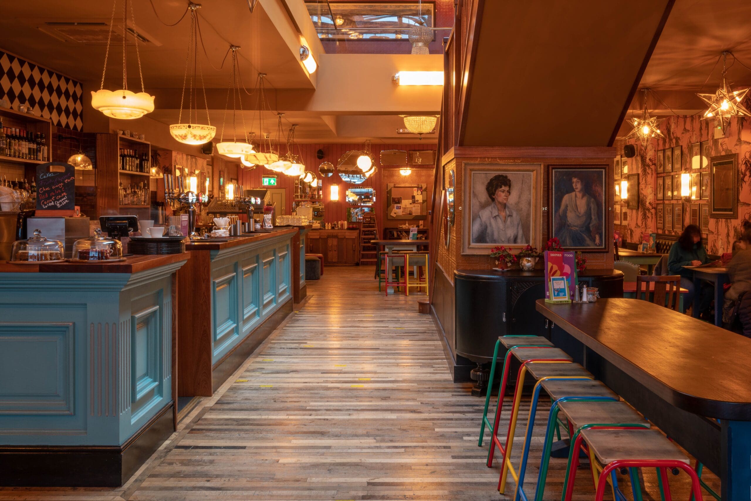 A cozy Portello bar interior with a blue counter on the left, colorful stools by dark tables on the right, warm lighting, framed paintings on the walls, and wooden floors. The space feels inviting and lively.