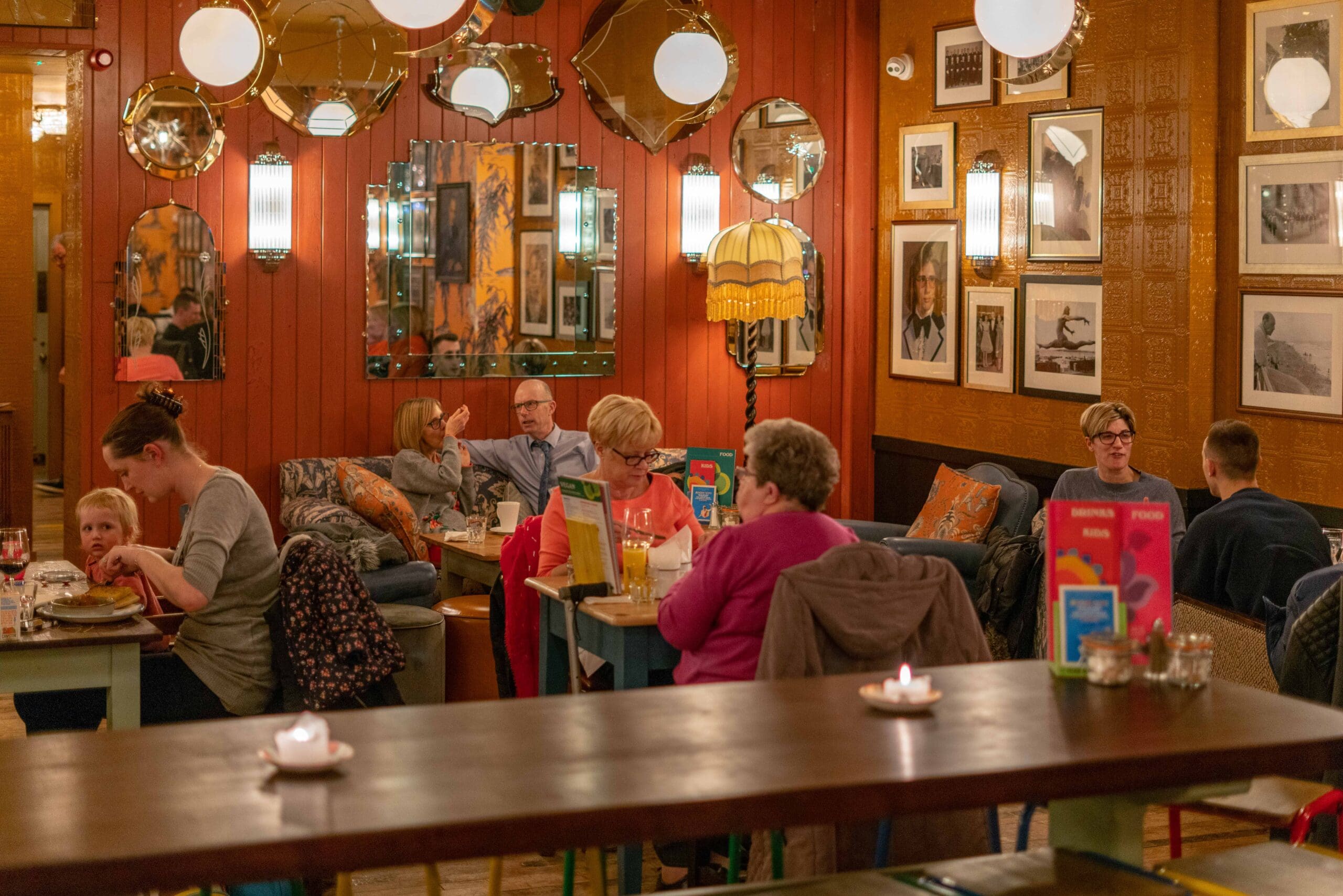 People of various ages sit and dine in a cozy, warmly lit restaurant with eclectic decor—framed pictures, mirrors, hanging lamps, and bottles of Portello. Guests enjoy eating, drinking, or talking at different tables.