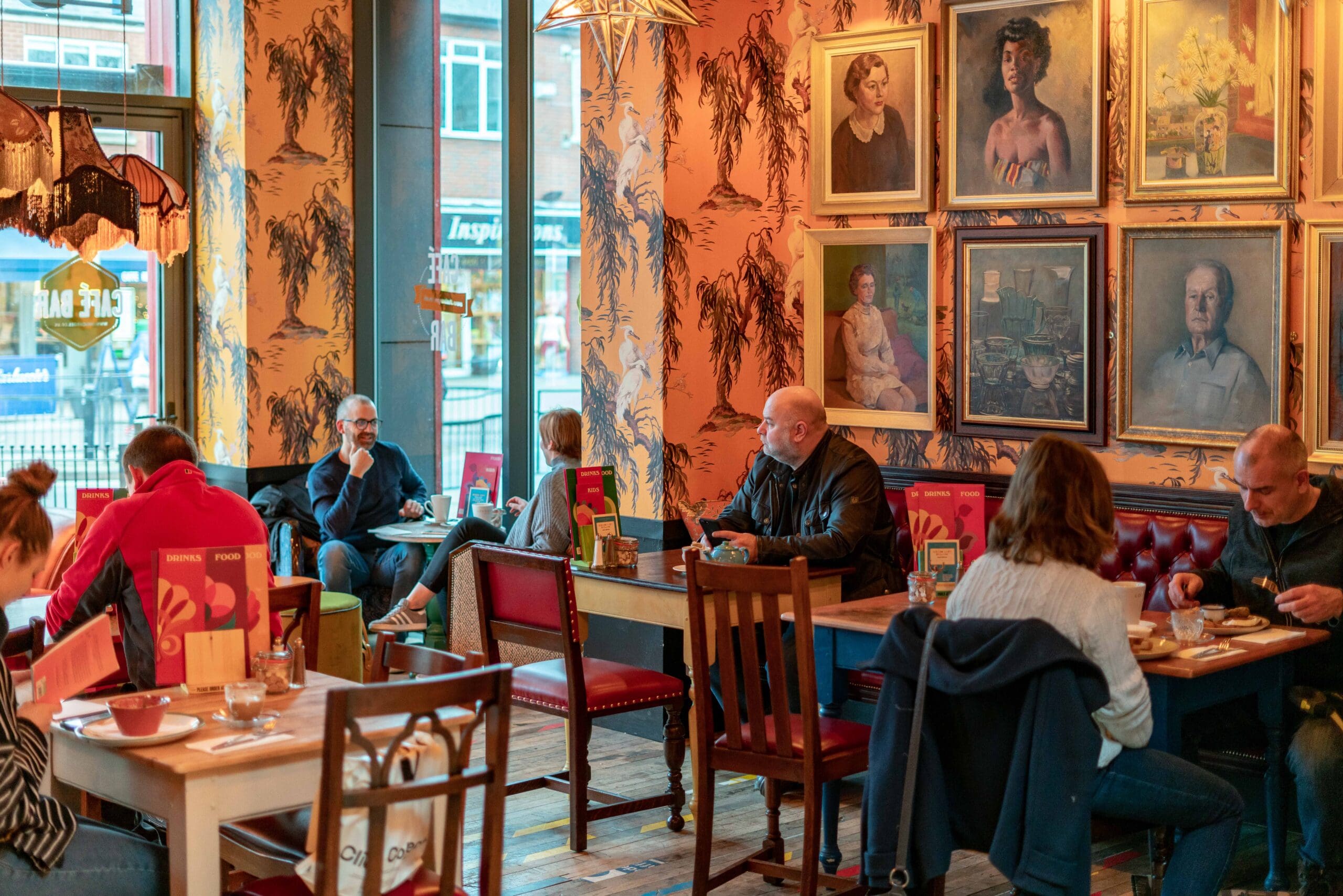 People sit at tables in Portello, a cozy, warmly lit cafe with vintage decor, orange patterned walls, framed portraits, and wooden furniture, enjoying food and conversation. Large windows let in natural light from outside.