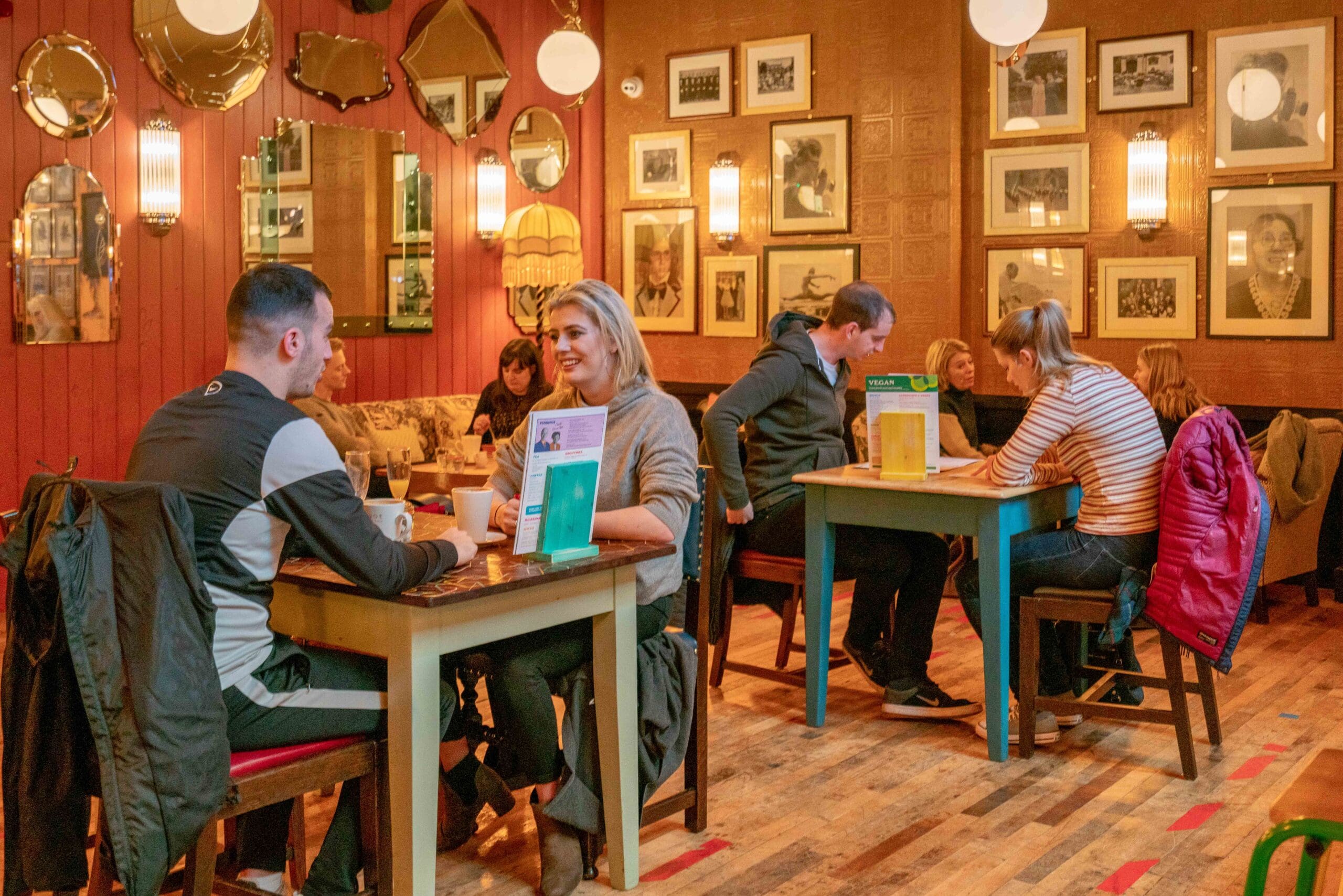 People sit at tables in a cozy, warmly-lit café with Portello bottles on the tables, framed photos and mirrors on the walls, chatting and enjoying drinks in a relaxed atmosphere.