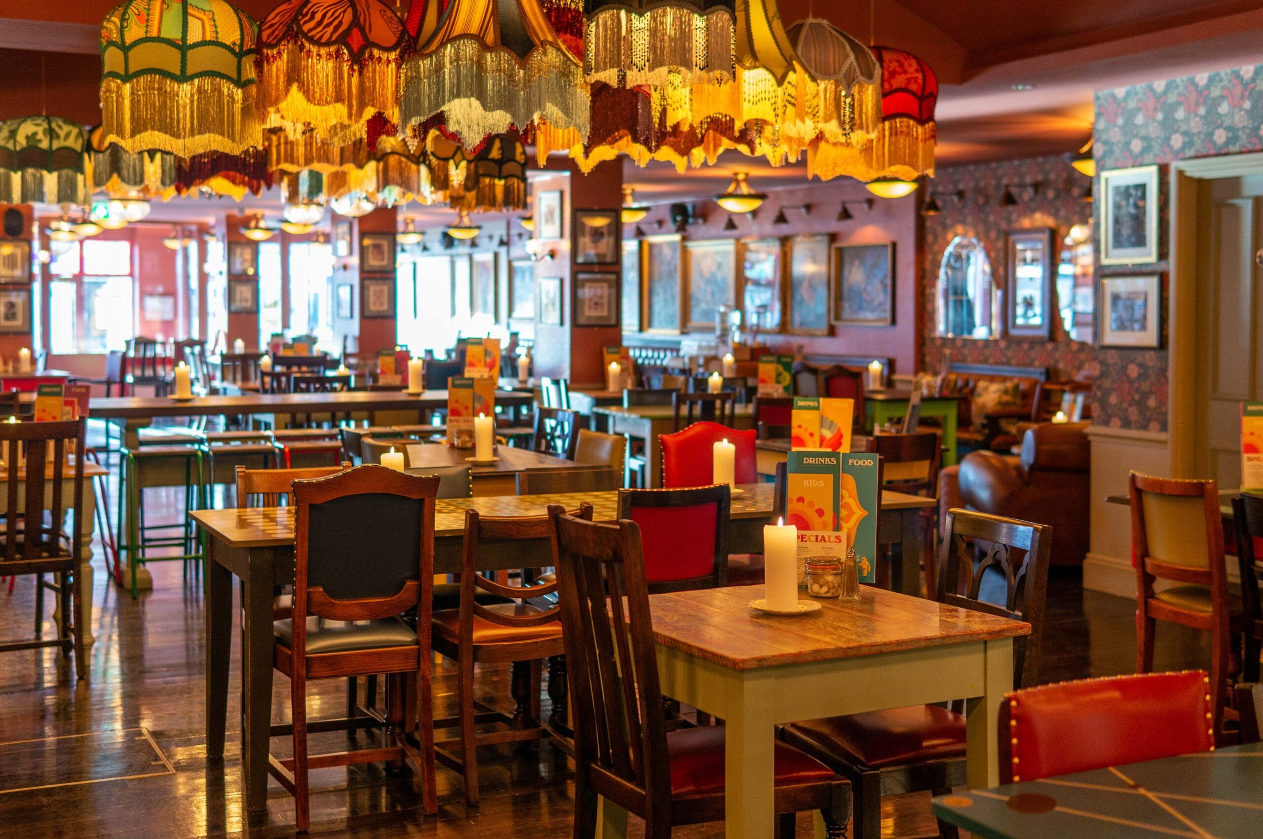 Colorful, eclectic restaurant interior with mismatched tables and chairs, Rosado vintage lampshades hanging from the ceiling, candles on tables, and framed art on the walls, creating a cozy and vibrant atmosphere.