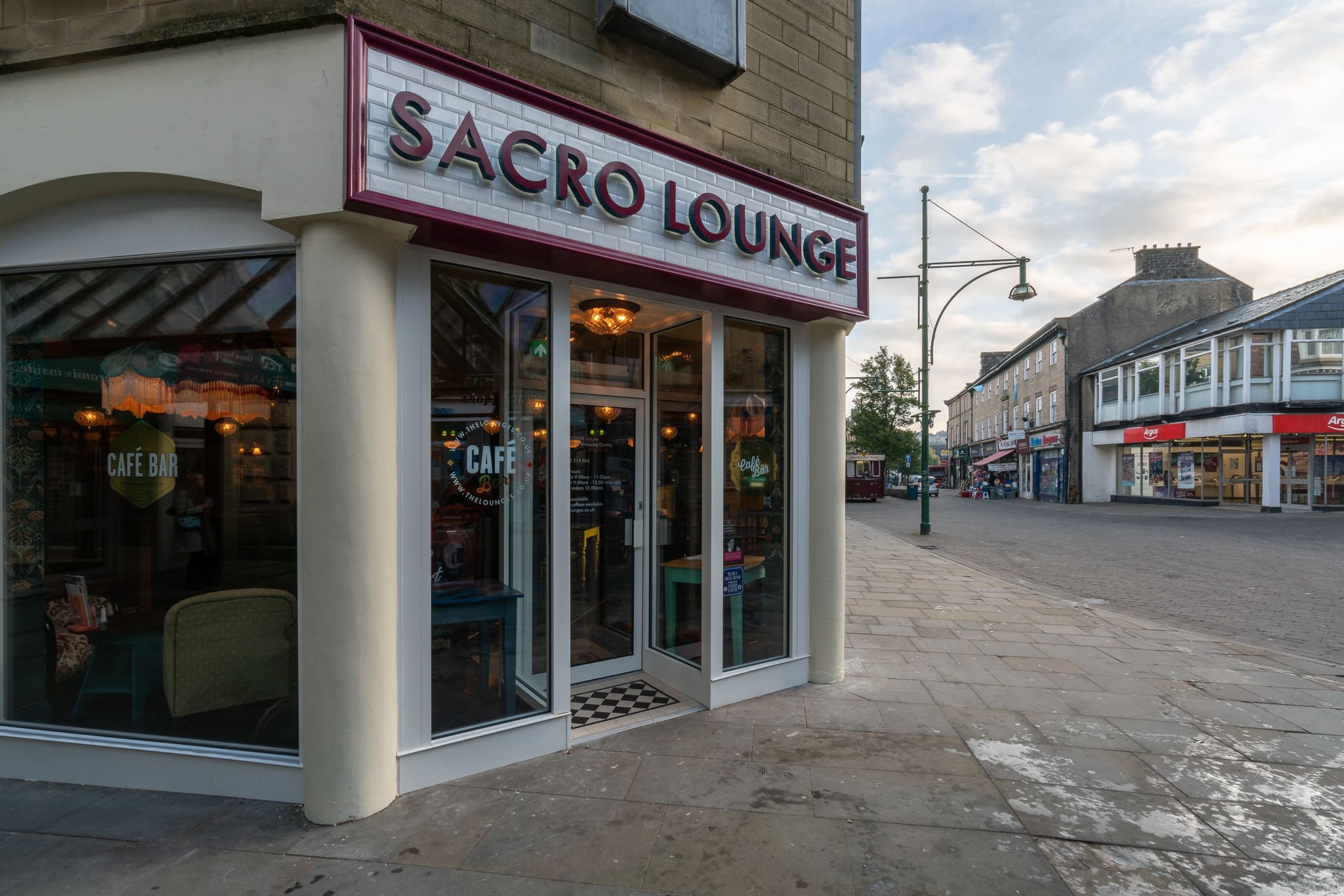 A street view of Sacro café bar with large windows and a glass door, located on a quiet sidewalk. Other shops line the street under a partly cloudy sky, highlighting the inviting atmosphere of Sacro.
