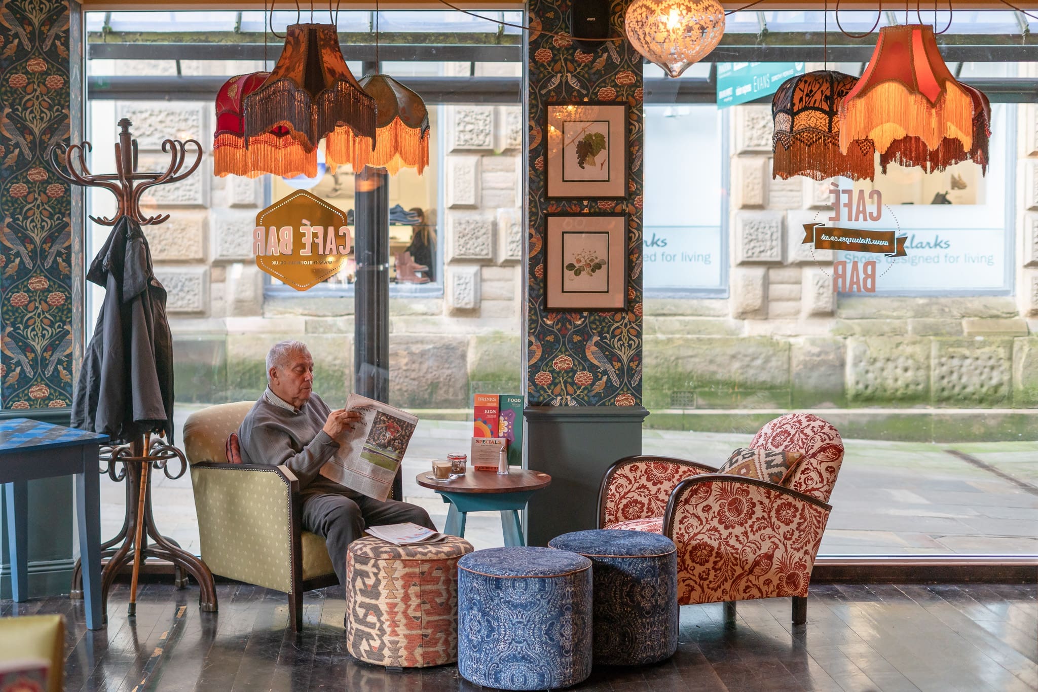 An older man sits in a cozy, vintage-style café reading a newspaper. The sacro ambiance is enhanced by ornate lamps, patterned wallpaper, colorful armchairs, and large windows looking out onto a stone building and street.