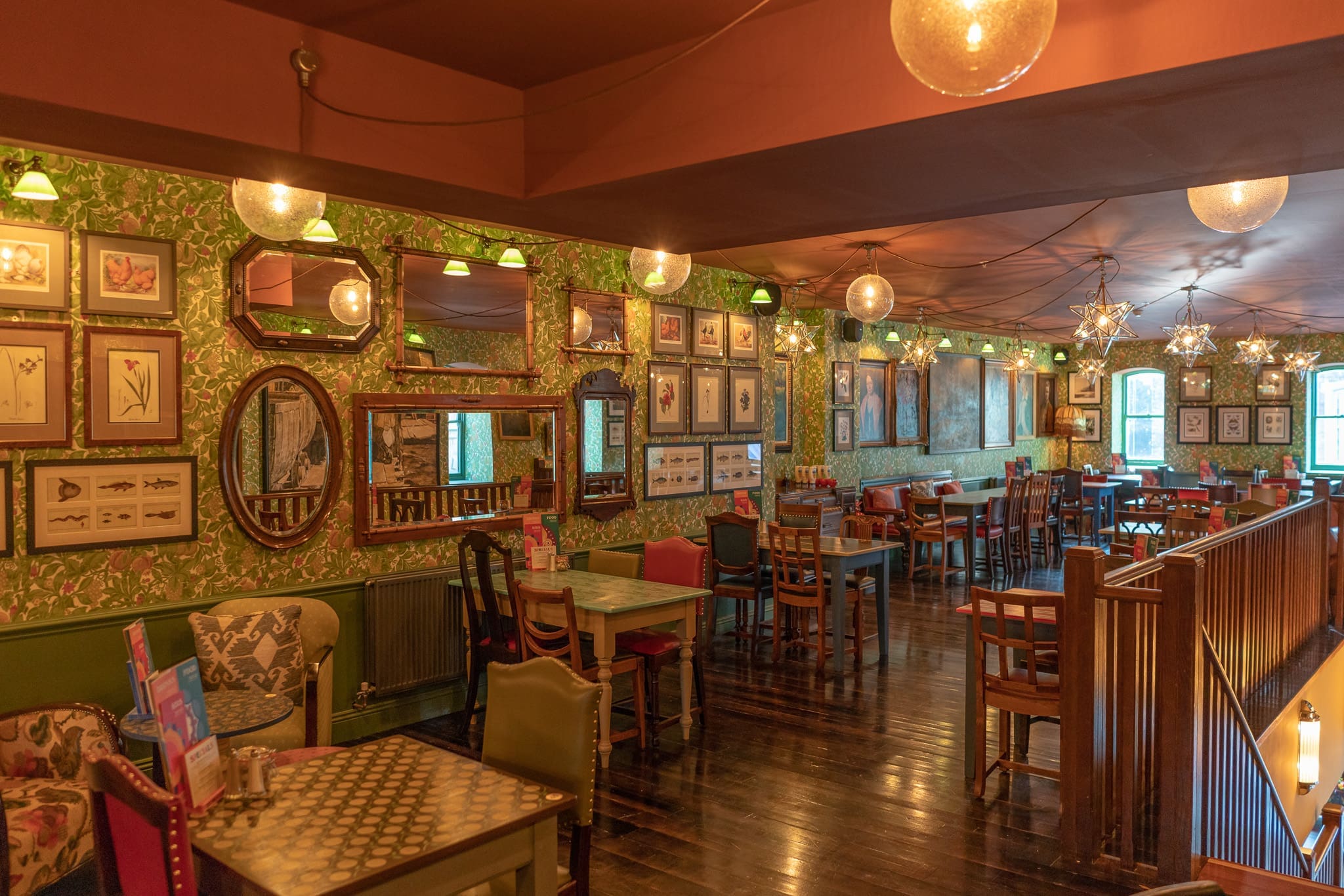 A cozy cafe with eclectic vintage decor and Sacro-inspired touches, green patterned wallpaper, framed art, and mirrors on the walls. Wooden tables and mismatched chairs fill the warmly lit room, creating an inviting atmosphere.