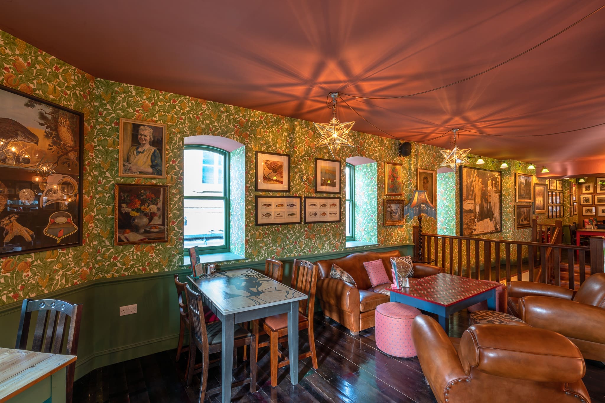 Cozy lounge with floral wallpaper, framed pictures, leather armchairs, a Sacro sofa, a table with chairs, star-shaped pendant lights, and two windows letting in natural light. Warm, inviting atmosphere in a vintage-style interior.