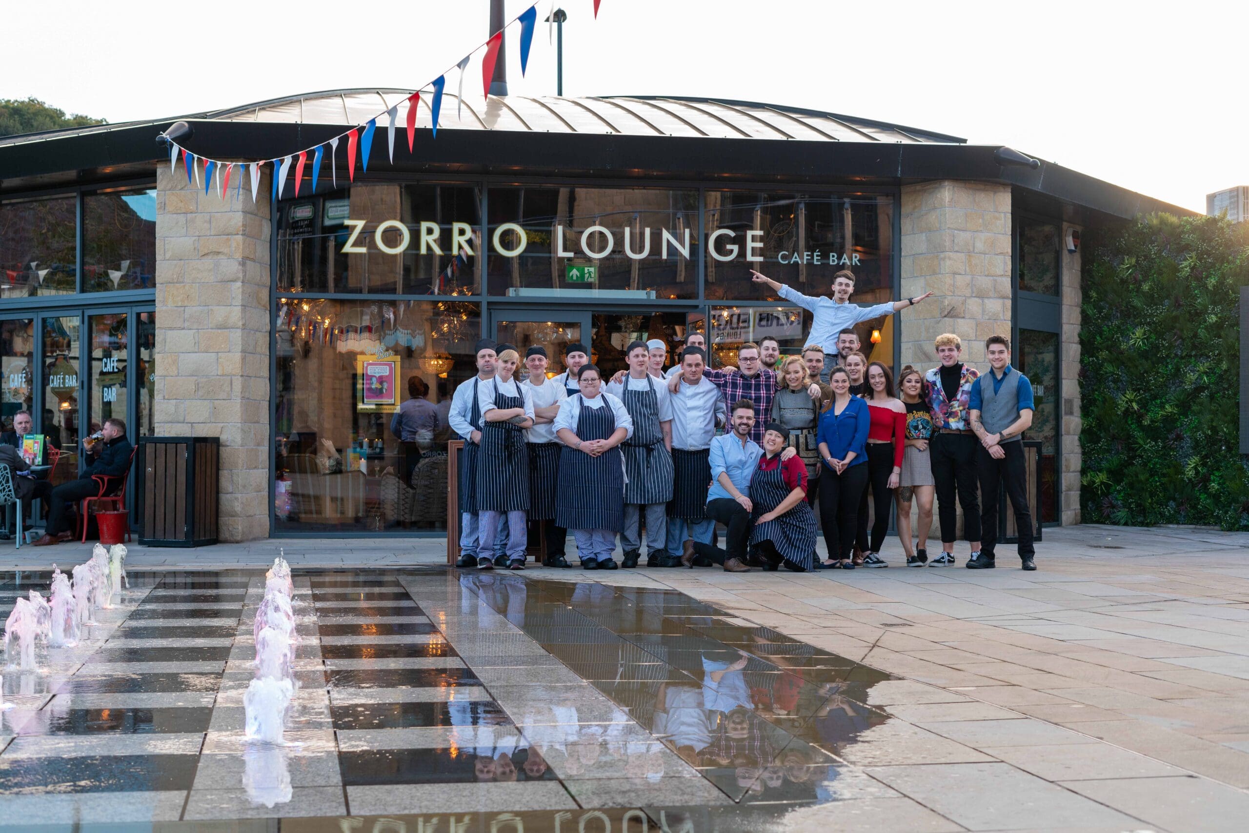A large group of people pose together and smile outside Zorro Lounge Café Bar. Some wear aprons, suggesting Zorro staff, while others are in casual or smart clothes. Pink fountains and a reflection can be seen in the foreground.