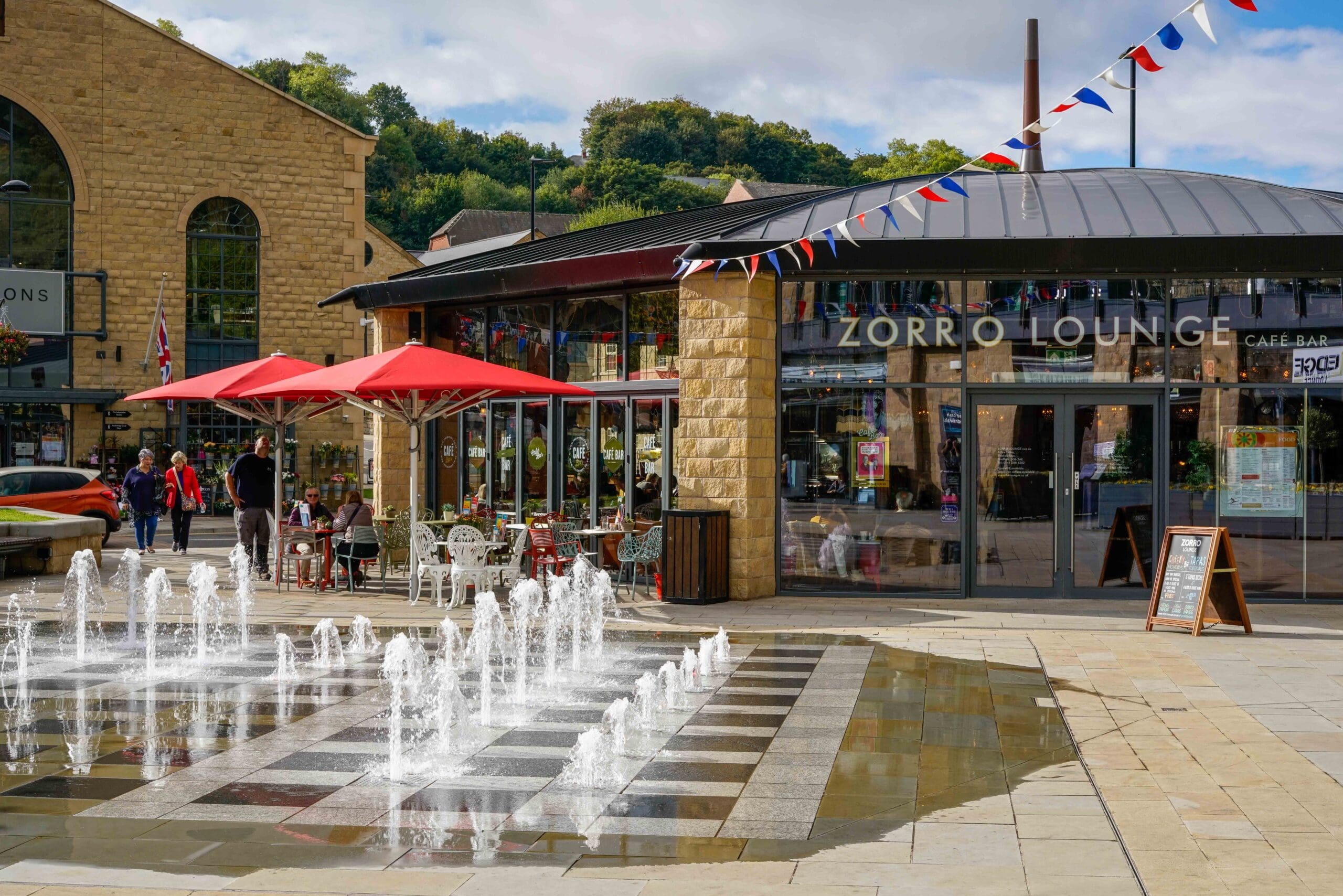 Outdoor scene featuring Zorro Lounge, with glass windows and red umbrellas shading outdoor tables. A decorative fountain with water jets sits in front, while colorful bunting hangs overhead and people stroll near the vibrant Zorro café.