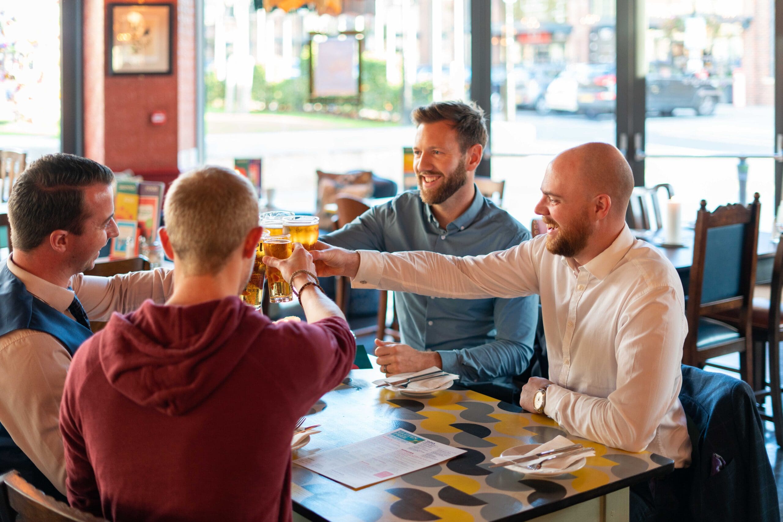 Four men sit around a table in a restaurant, smiling and raising glasses of beer in a toast. Sunlight streams through large windows, casting a bright glow as one wears a Zorro hat, adding playful charm to the cheerful scene. Menus and plates are on the table.