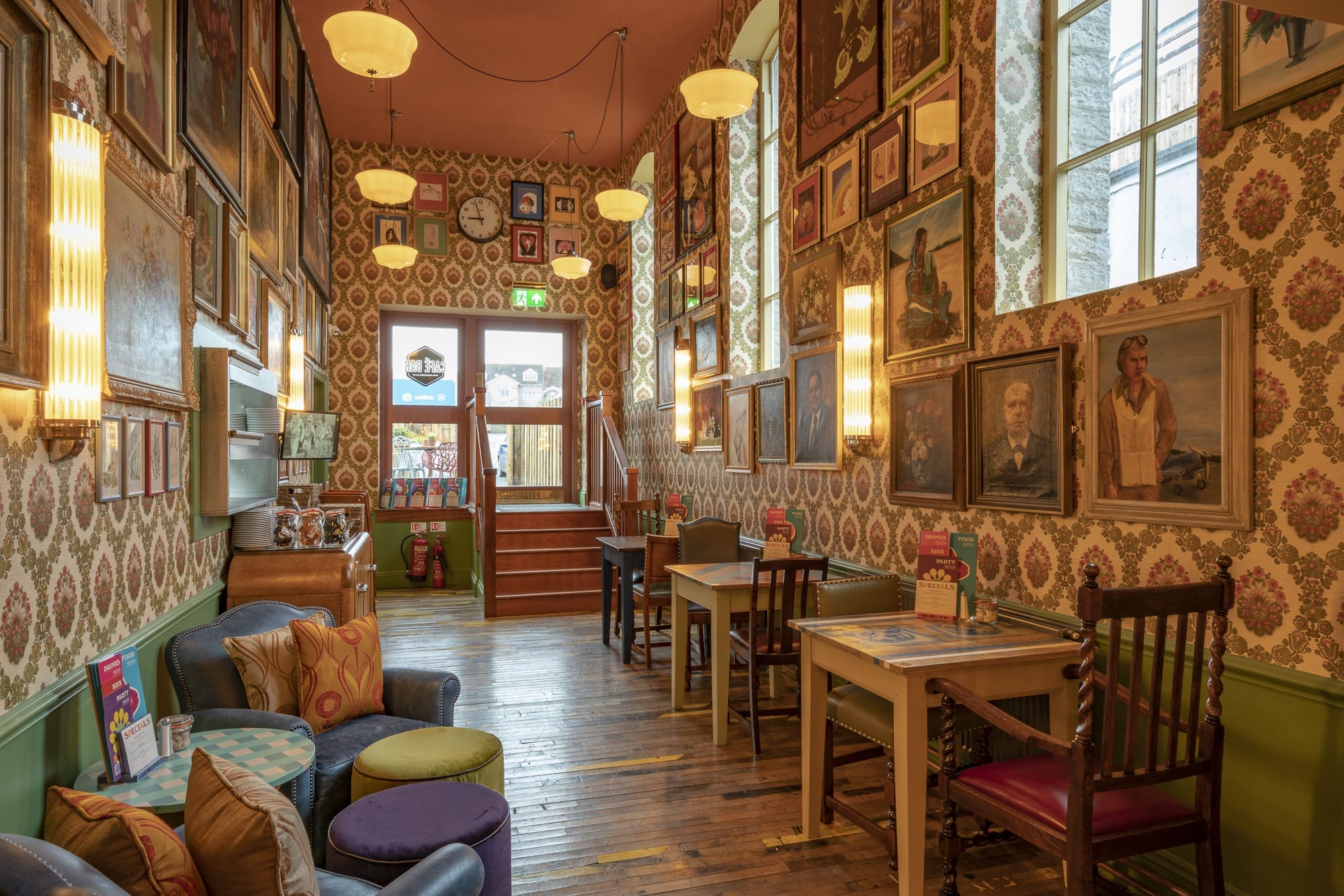 A cozy vintage cafe with colorful armchairs and wooden tables, set against a fondo of patterned wallpaper, hanging lights, framed portraits, and tall windows letting in natural light. The atmosphere is warm and inviting.