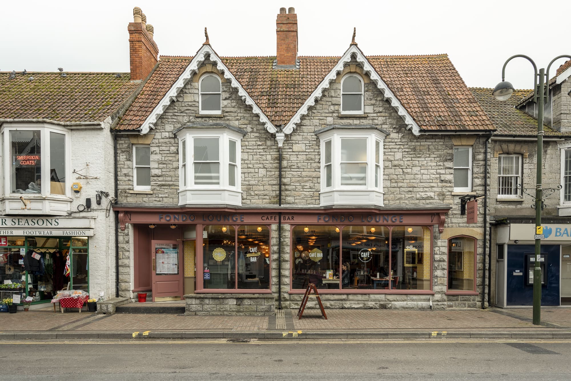 A stone-fronted building with two gables houses "Pondo Lounge" café bar on a street. Shopfronts with large windows display warm lighting. Neighboring stores include Fondo Florist on the left and a bank on the right.