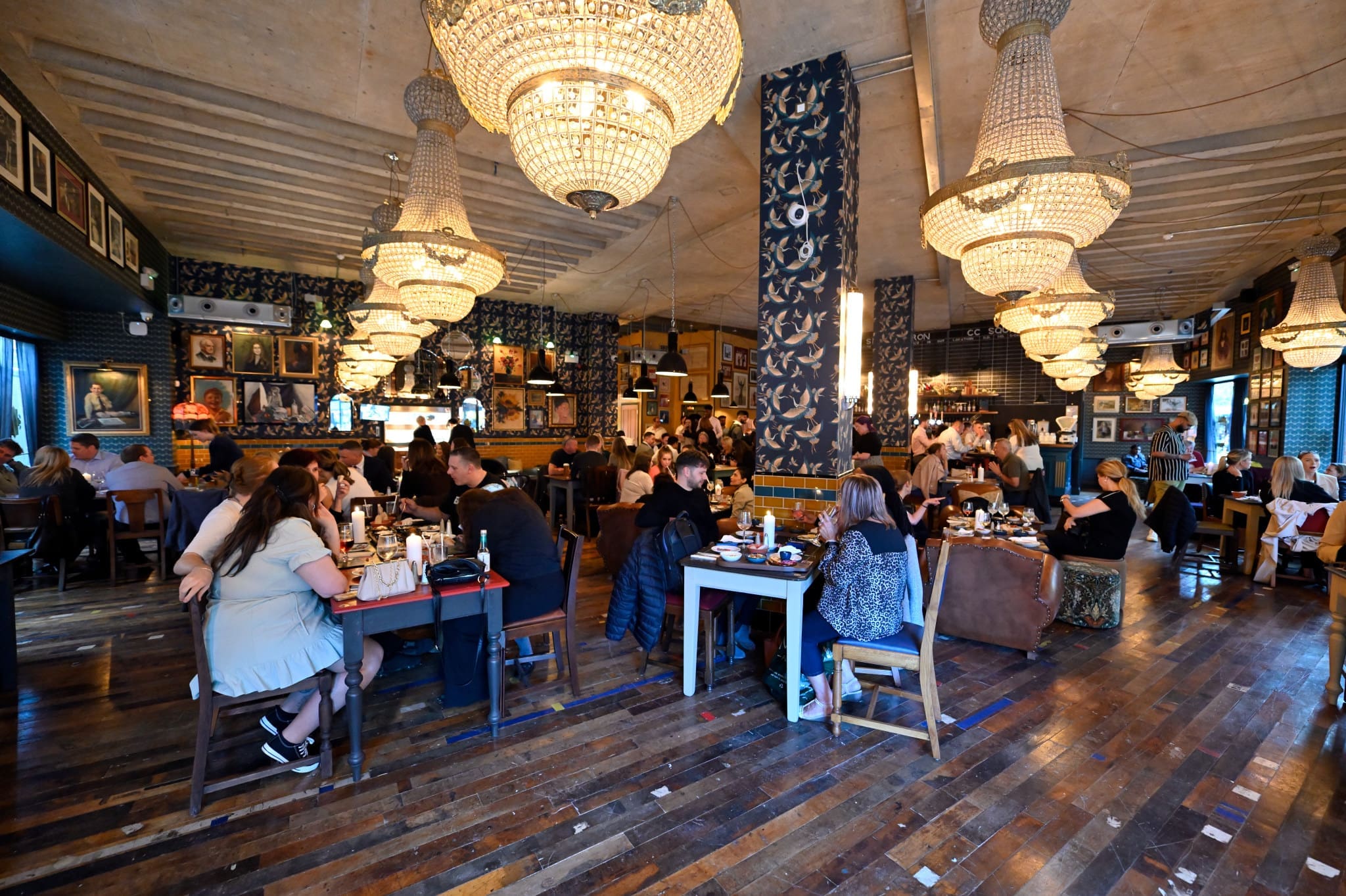 A lively restaurant with large chandeliers, people dining at various tables, wooden floors, and eclectic wall decor featuring framed pictures and patterned wallpaper. The warm, bustling atmosphere is enhanced by signature Morello-inspired dishes.